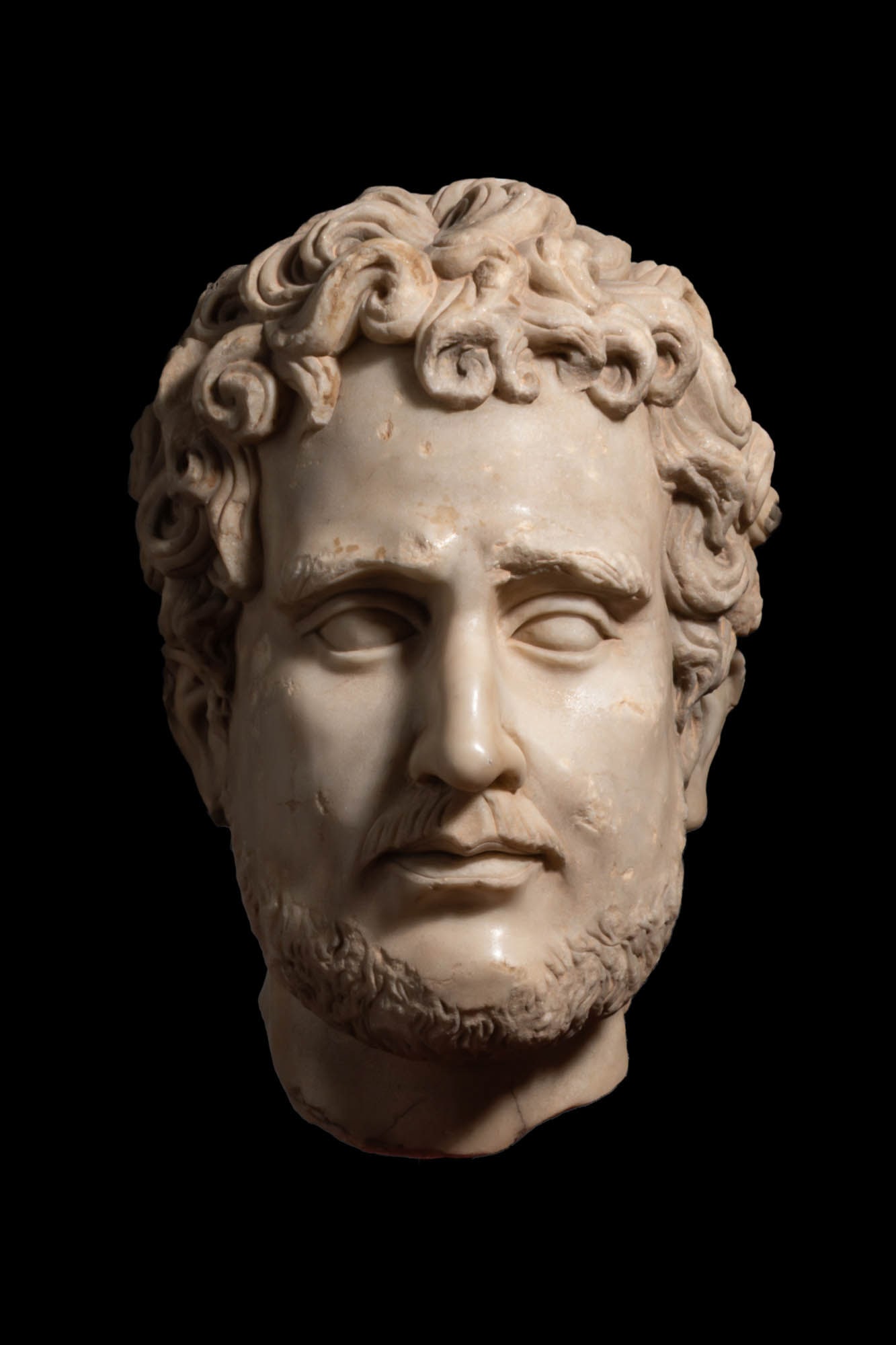 ROMAN MARBLE HEAD OF ANTONINUS PIUS: Ca. AD 138 - 161 A marble portrait head of Antoninus Pius, with a full crown of deeply drilled curls arranged in thick locks over the forehead and temples. Heavy brows sit above almond-shaped eyes, th
