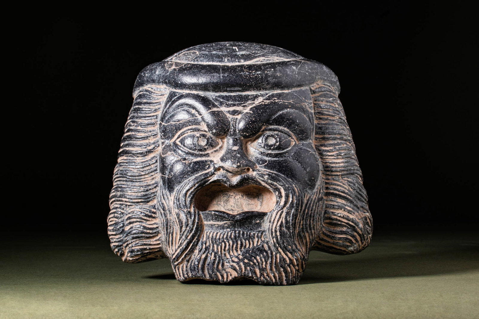 RARE ROMAN BLACK STONE THEATRE MASK: Ca. AD 100. An impressive black stone theatre mask with realistically rendered facial features. It has a coiffure featuring curled bangs with long ringlets falling to either side of the chin, a long m