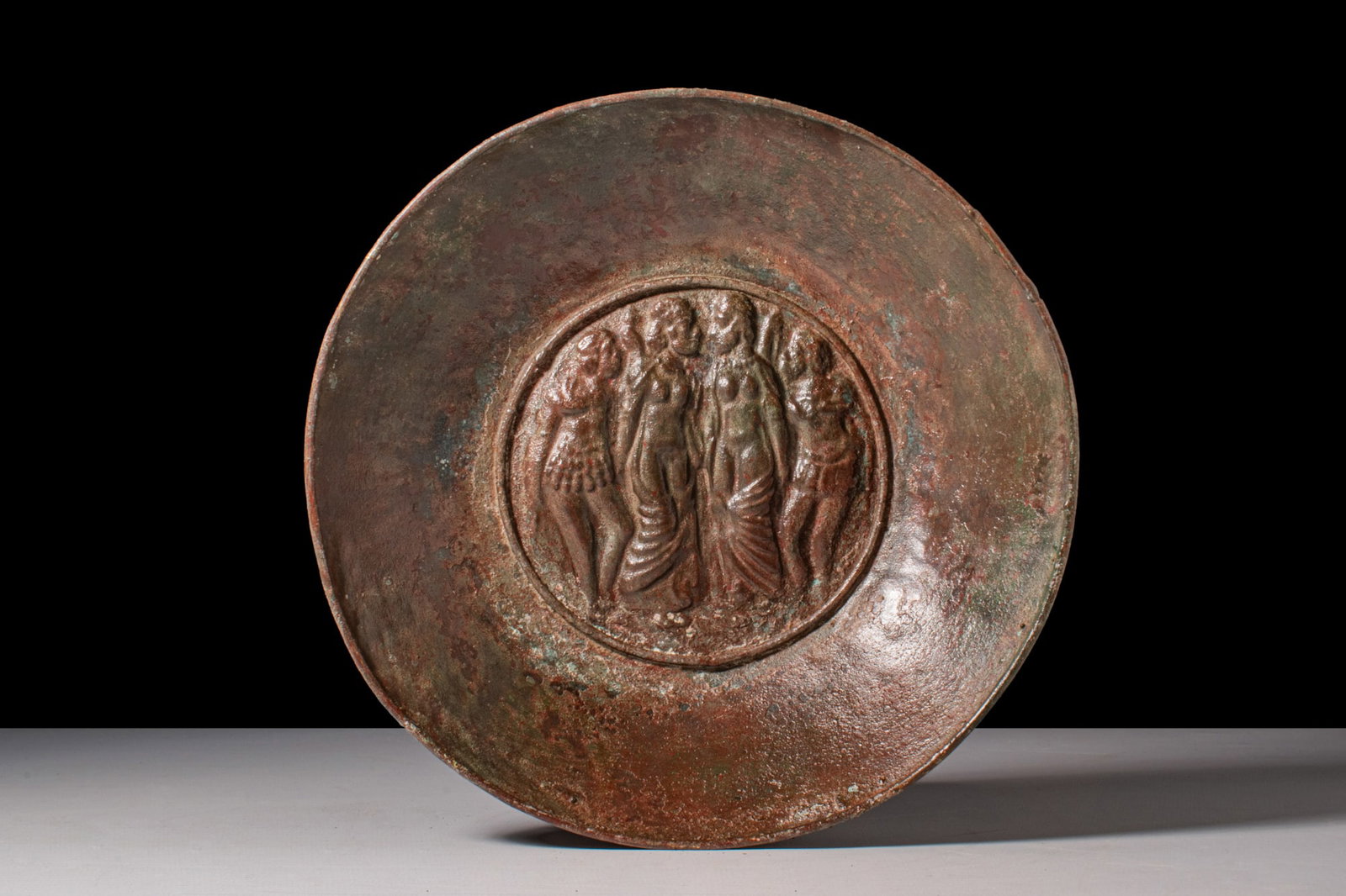 ROMAN BRONZE DISH WITH HAMMERED DETIES: Ca. AD 150A bronze Eatern Roman of Gandharan dish formed from a hammered sheet with a broad, shallow wall and a rounded rim, the interior centred by a raised circular medallion in high relief with fou
