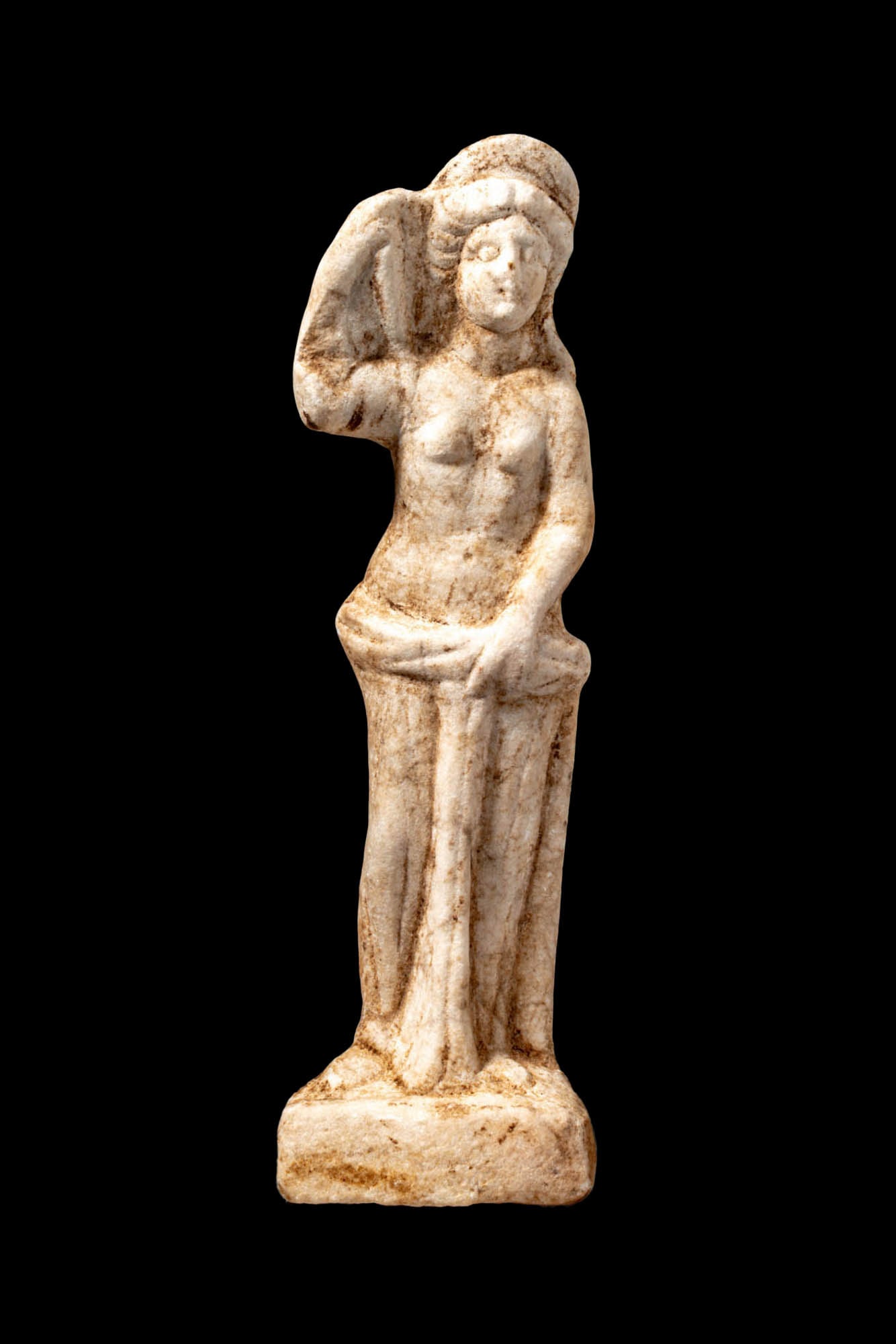 ROMAN MARBLE STATUETTE OF BATHING VENUS: Ca. AD 100 - 300. A marble figure of bathing Venus depicted nude, standing on an integral flat plinth. Her right arm is raised to touch the back of her head, while her left hand grasps the drapery wra