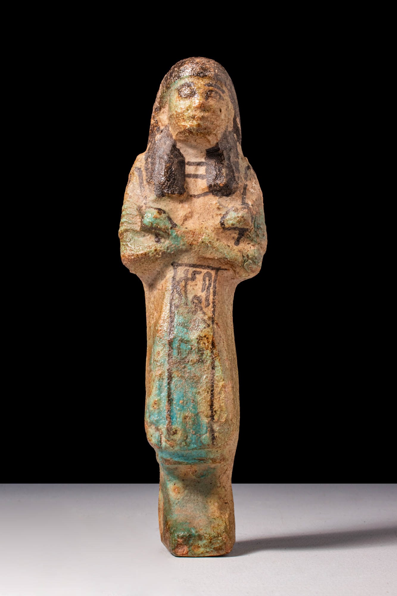 EGYPTIAN FAIENCE USHABTI OF ANONYMOUS: New Kingdom, XIX - XX Dynasty, Ca. 1292 - 1069 BC. A faience ushabti figure with arms crossed over the chest, the hands clutching agricultural implements. The face is modelled with full cheeks and a