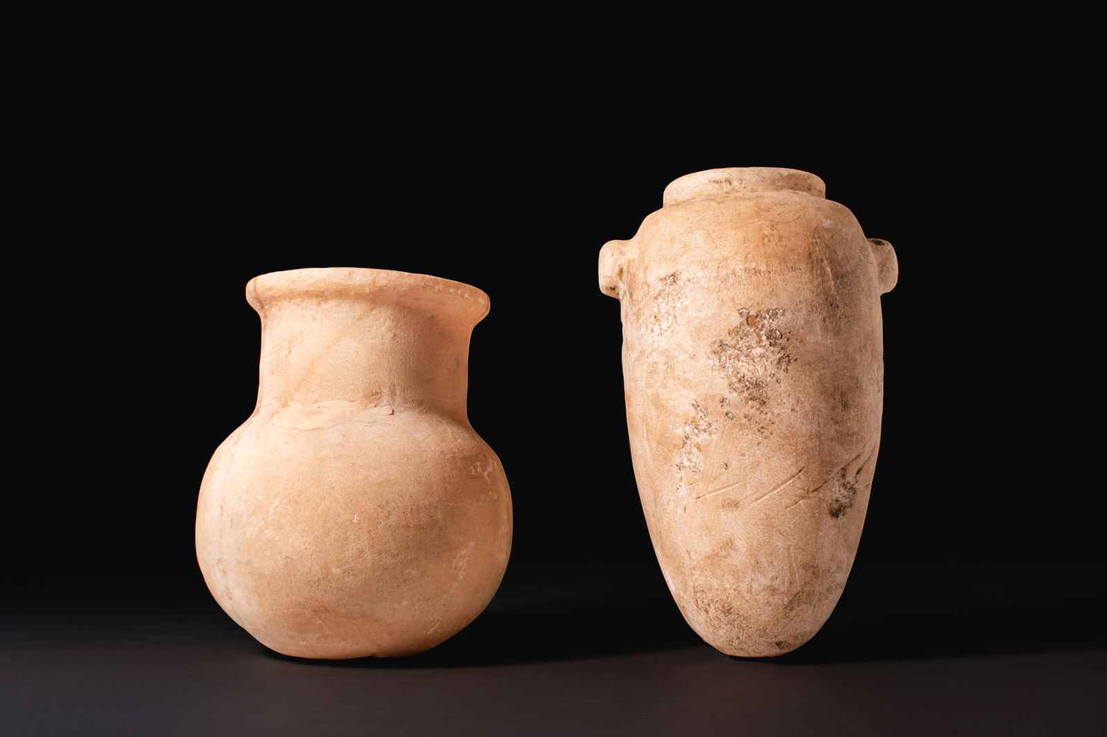 PAIR OF EGYPTIAN STONE VESSELS: Old Kingdom, Ca. 2640 - 2200 BCA pair of alabaster vessels, including, from left to right: a squat globular jar with a rounded body rising to a broad neck ending in a thick, pronounced rim; and a