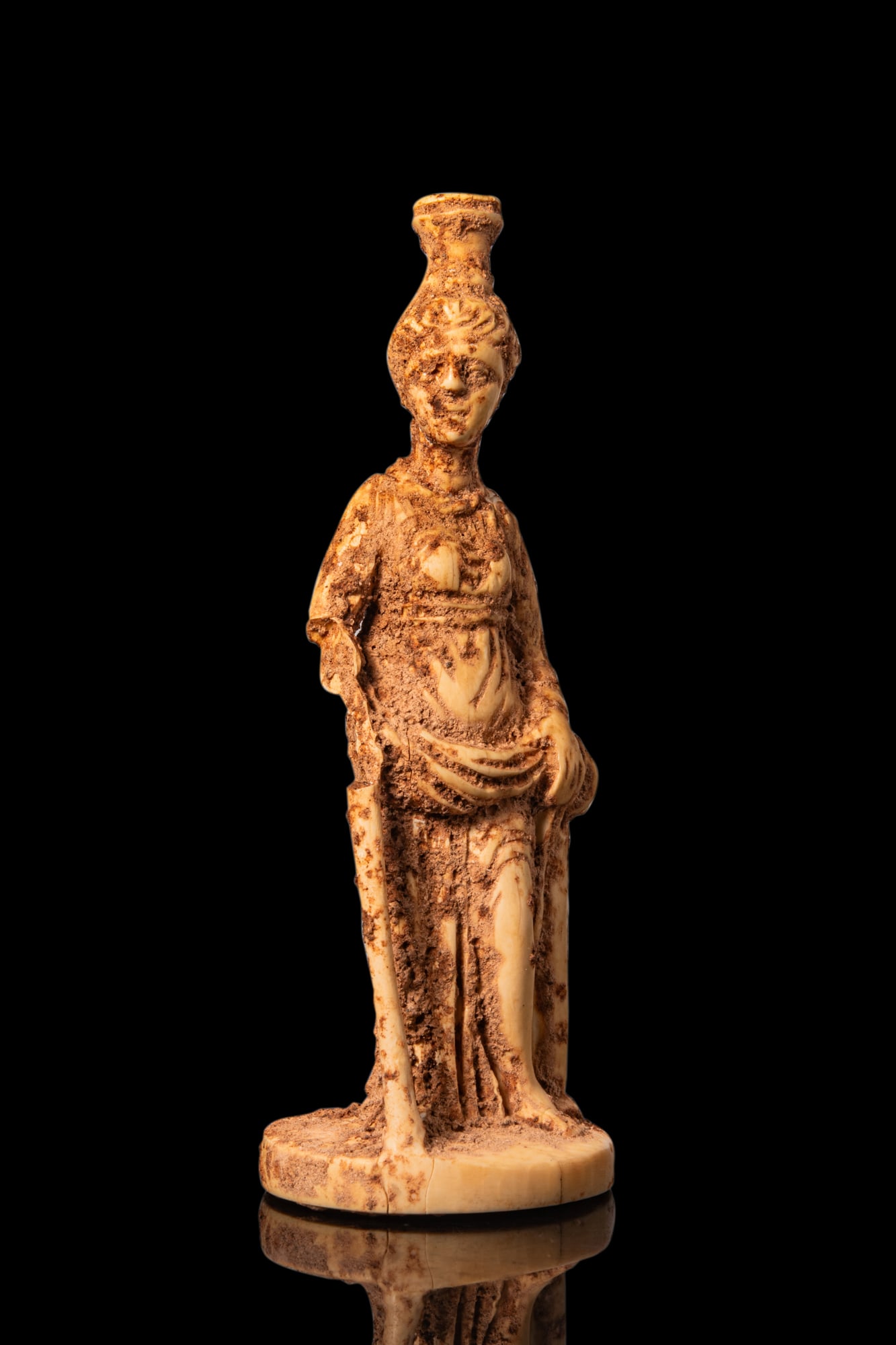 ROMAN BONE FIGURINE OF ISIS-FORTUNA: Ca. AD 1 - 300A bone figurine of Isis standing on an integral circular base, wearing a long belted robe falling to the feet. She lifts the front hem to reveal the left leg. Along her right side is the