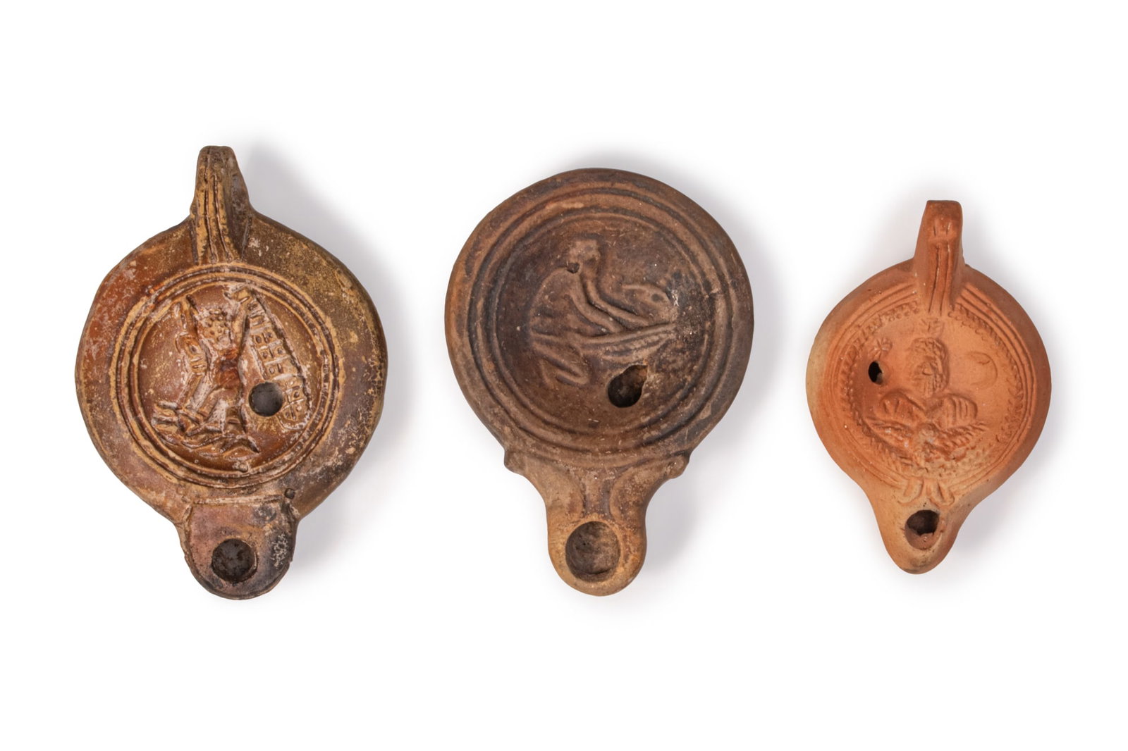 COLLECTION OF ROMAN TERRACOTTA OIL LAMPS: Ca. AD 1 - 300A collection of three mould-made terracotta oil lamps, including, from left to right: a lamp with rounded body, rear handle and short nozzle, the circular discus framed by double
