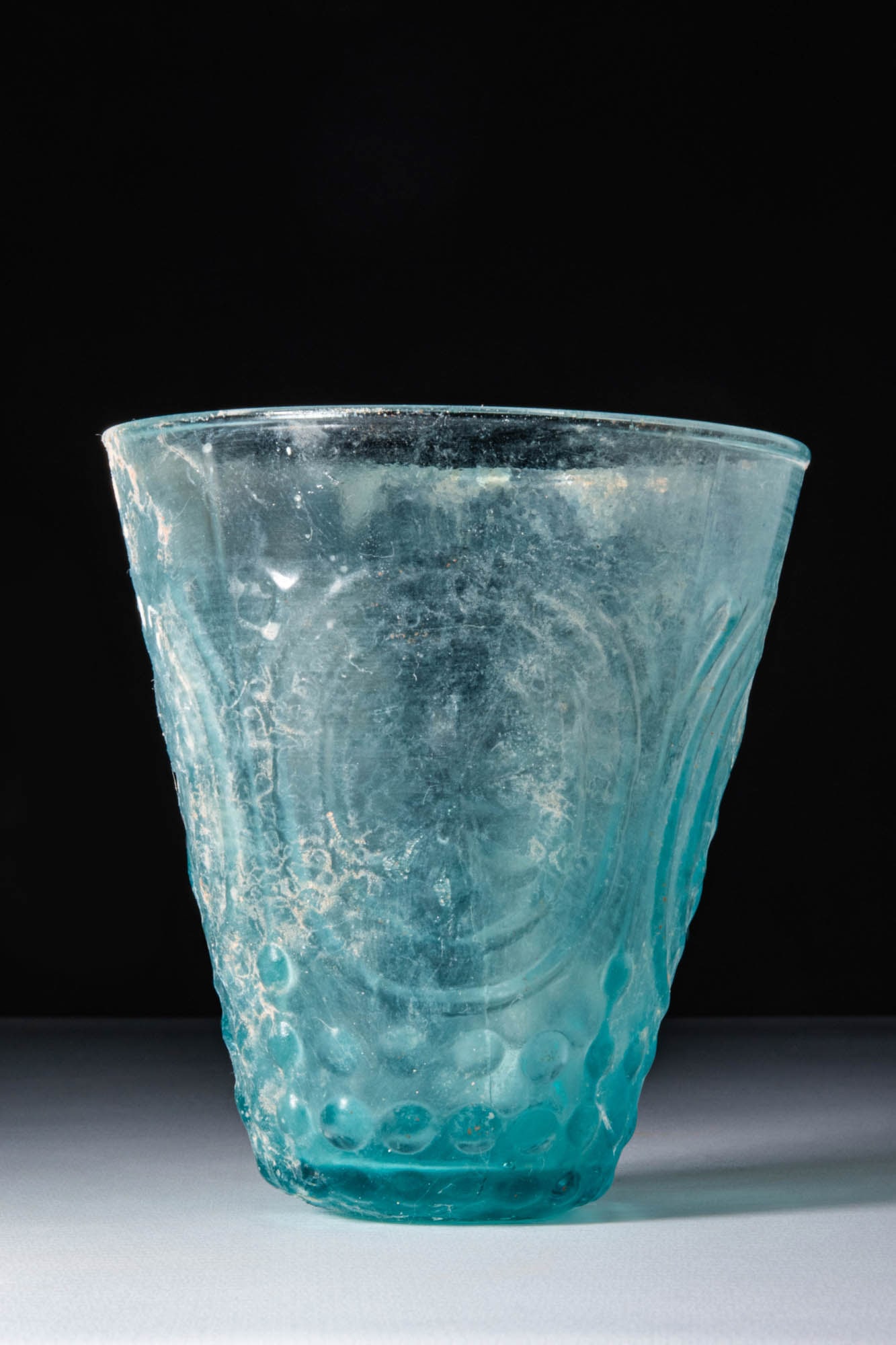 ROMAN BLUE GLASS BEAKER: Ca. AD 1 - 50. A pale blue glass beaker with a flaring rim and conical body. The surface is decorated with a pattern of large oval medallions surrounded by concentric bands and raised circular bosses