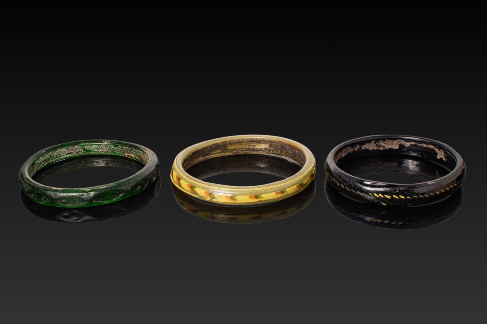 COLLECTION OF ROMAN POLYCHROME AND MONOCHROME GLASS BANGLES: Ca. AD 1 - 300A collection of three glass bangles, including, from left to right: a dark green monochrome bangle; a yellow bangle with a raised outer rib formed by an applied trail of opaque yellow,