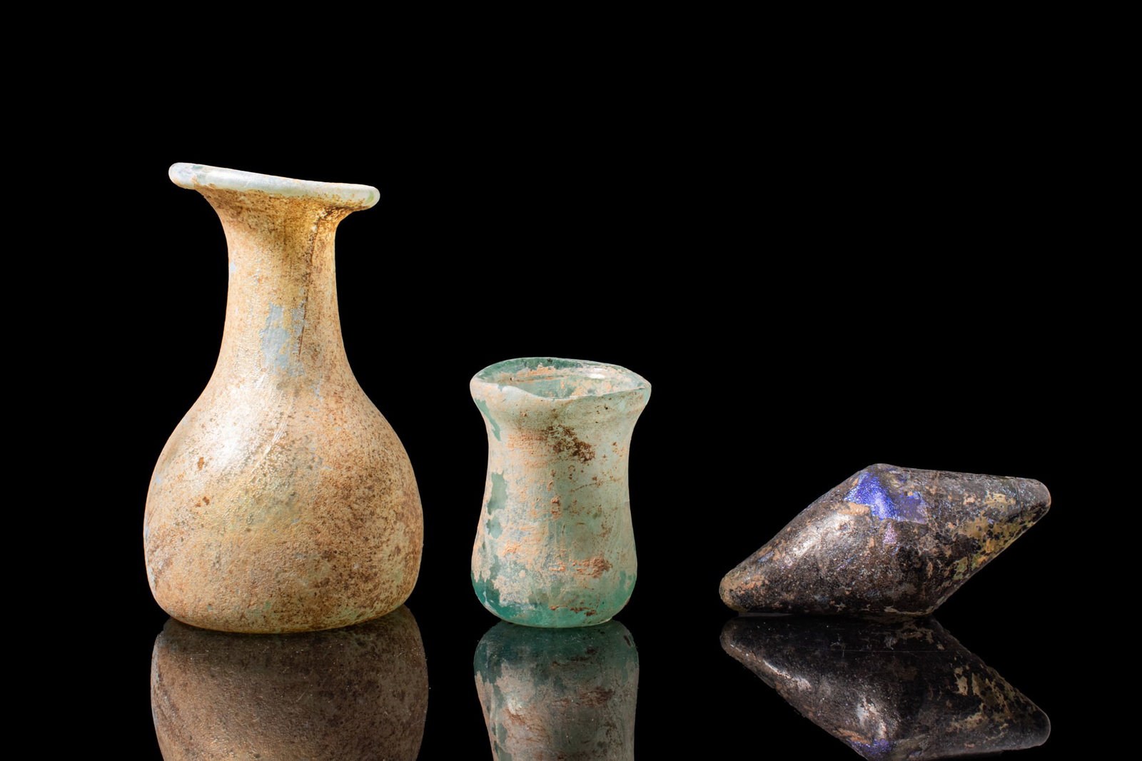 ROMAN GLASS BOTTLE, CUP AND BICONICAL STOPPER: Ca. AD 200 - 400A collection of three glass objects, including, from left to right: a pale blue bottle with a bell-shaped body narrowing to a tall neck and a funnel mouth with an outturned rim; a