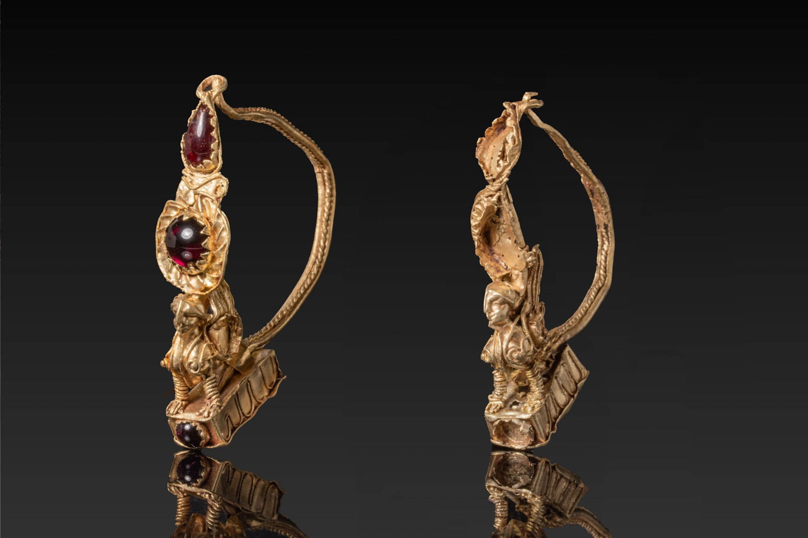PAIR OF GREEK HIGH KARAT GOLD AND GARNET EARRINGS WITH SPHINXES: Ca. 200 - 1 BC. A matched pair of gold earrings, each with a winged sphinx seated on a rectangular plinth with a frieze of tongues. The creature is modelled with a female head, the body adorned with c