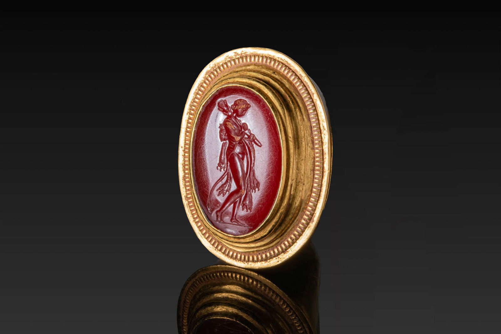 LARGE HELLENISTIC HIGH KARAT GOLD RING WITH CARNELIAN INTAGLIO OF OMPHALE: Ca. 200 - 1 BC. A large gold ring with a broad, tapering hoop expanding to an oval bezel plate, the mount formed with stepped collars and an outer beaded border. Set with an oval carnelian intaglio en