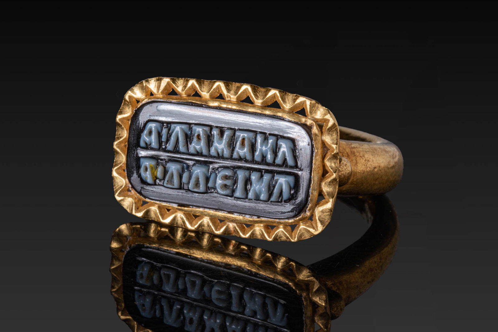 ROMAN HIGH KARAT GOLD OPENWORK RING WITH INSCRIBED SARDONYX: Ca. AD 300. A gold ring with a rounded hoop supporting a rectangular bezel plate, framed by an openwork radiating border. The bezel is set with a sardonyx plaque with a two-line inscription. Size: D:1
