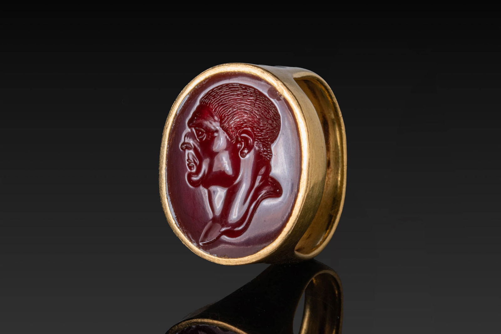 ROMAN HIGH KARAT GOLD RING WITH CARNELIAN INTAGLIO PORTRAIT OF EMPEROR TITUS: Ca. 100 BC - AD 100. A gold ring with a broad, flat-section hoop widening to an oval bezel with a plain frame securing a carnelian intaglio. The stone is engraved with the profile portrait of emperor