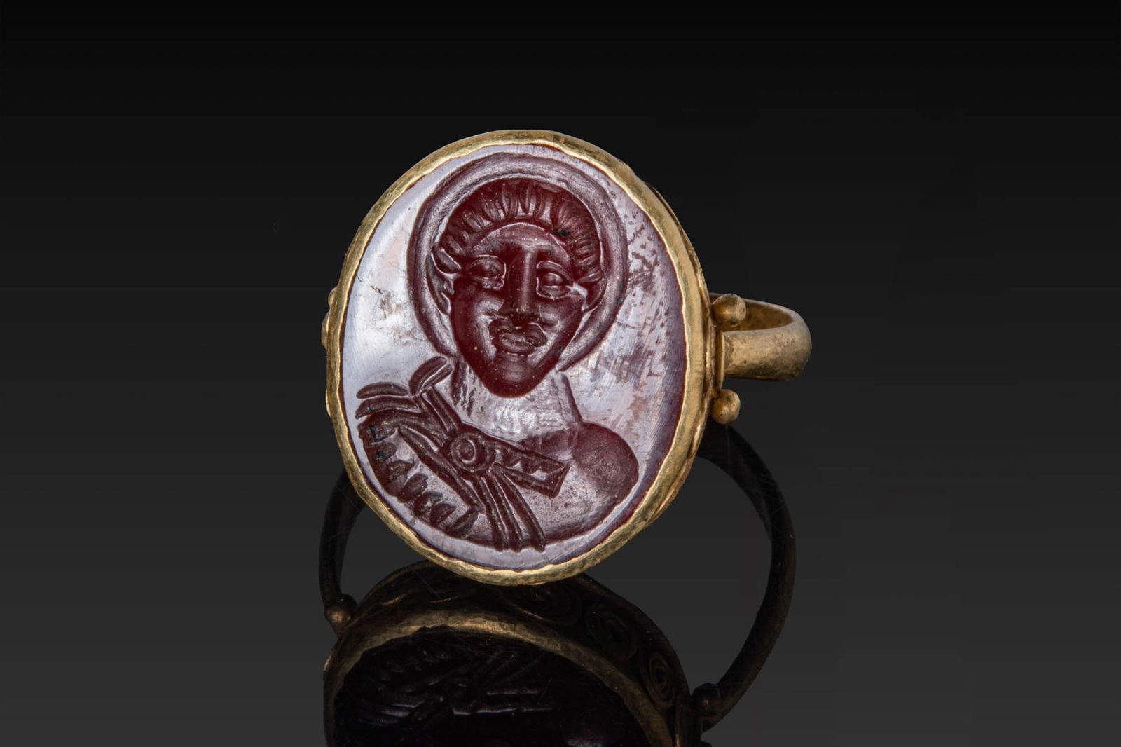 BYZANTINE CARNELIAN INTAGLIO OF A MILITARY SAINT IN HIGH KARAT GOLD RING: Ca. AD 900 - 1200. A carnelian intaglio engraved with a bust of a haloed saint with a short moustache and the hair arranged in close rows, the garment tied by a circular brooch. Mounted in a possibly