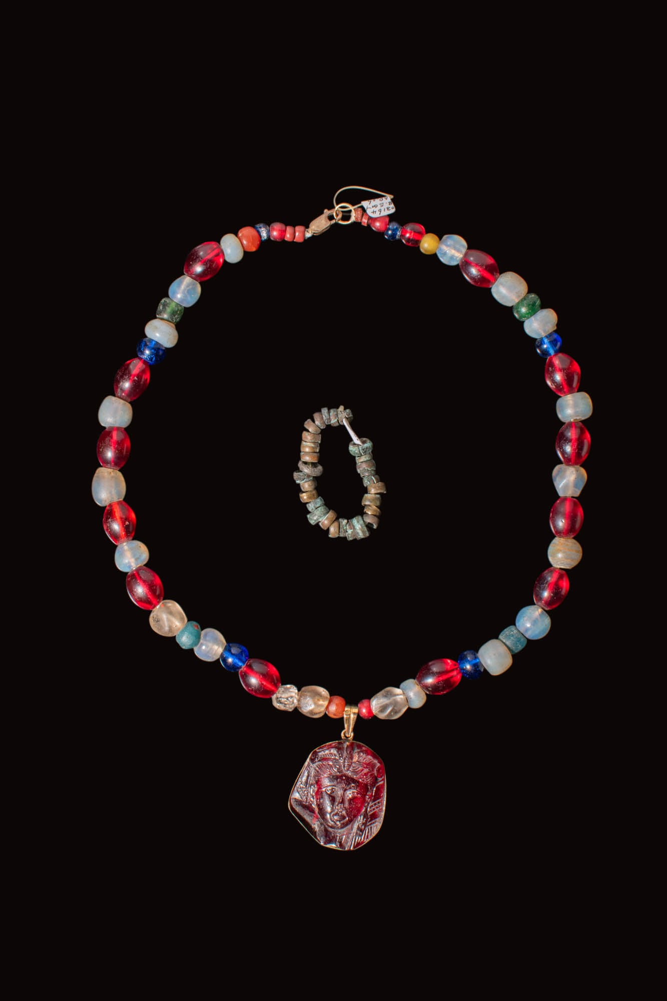 STUNNING GRAECO-EGYPTIAN STONE AND GLASS BEADED NECKLACE WITH PENDANT DEPICTING NOBLEWOMAN: Ca. 332 - 30 BC. A beaded necklace strung with alternating translucent and opaque glass and stone beads in red, beige and cobalt, with occasional green and amber. Suspended at the centre is a pendant