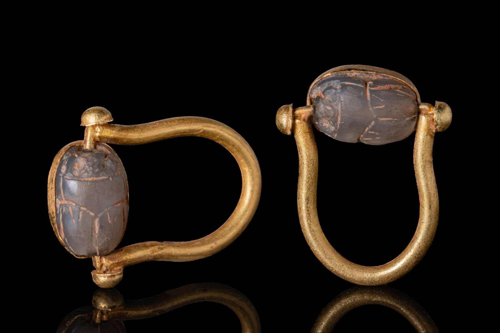 EGYPTIAN CHALCEDONY SCARAB IN HIGH KARAT GOLD SWIVEL RING: Ca. 500 - 300 BC. A chalcedony scarab with incised wing cases, pierced lengthwise, the underside plain. Mounted in a possibly later gold swivel ring with a thick, rounded hoop and an oval frame betwee