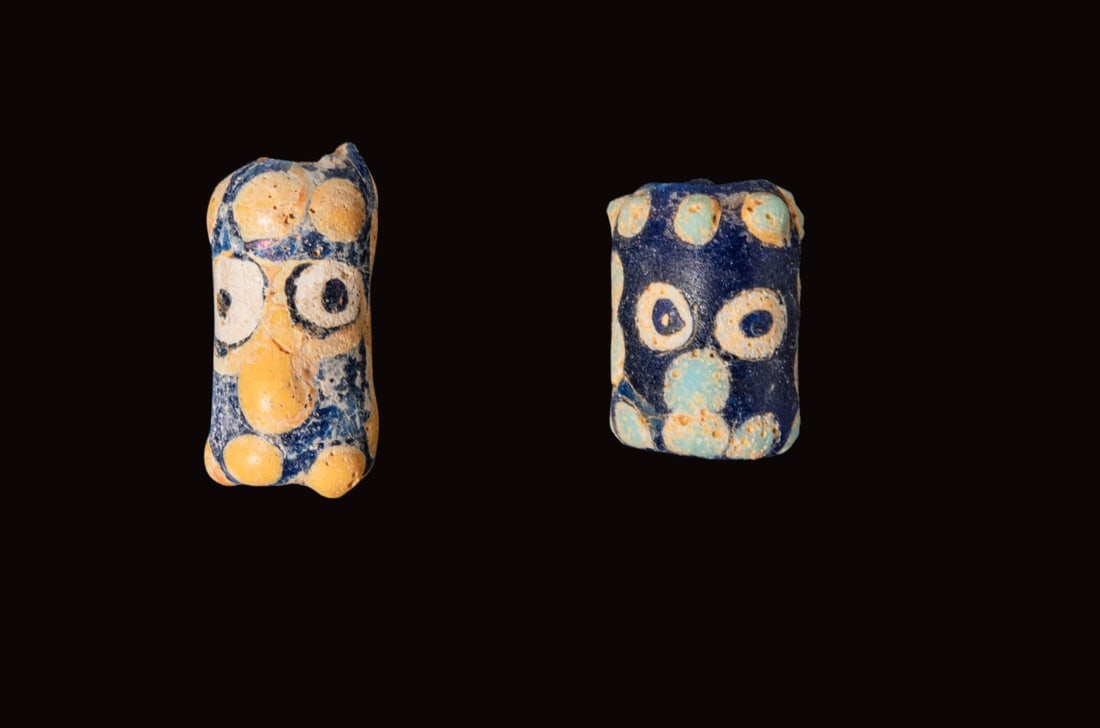 PAIR OF PHOENICIAN FACE BEADS: Ca. 500 - 300 BC. A pair of glass beads, from left to right: an elongated cylindrical bead with a stylised face in applied white, with round eyes and a long projecting nose, with horizontal rows of sm