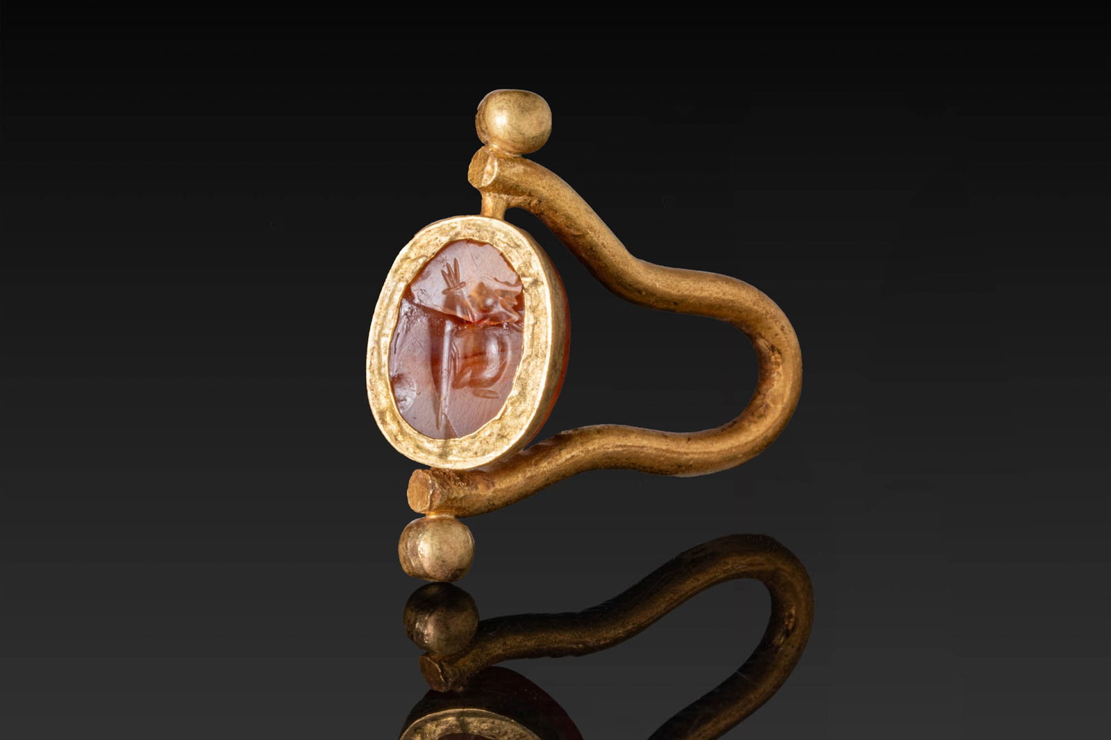 GREEK CARNELIAN SCARAB WITH HARE IN HIGH KARAT GOLD SWIVEL RING: Ca. 500 - 300 BC. A carnelian scarab, pierced lengthwise, engraved on the flat underside with a hare in profile. Mounted in a possibly later gold swivel ring with a looped hoop and a central oval fram