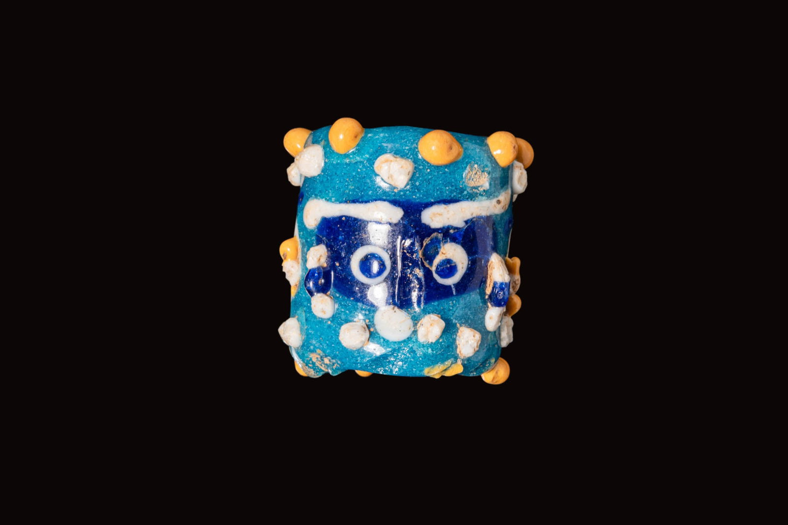 PHOENICIAN GLASS FACE BEAD: Ca. 500 - 300 BC. A blue glass bead of cylindrical form with three faces spaced around the circumference, each with a prominent white nose, white and yellow ears, and eyes with blue pupils, the remain
