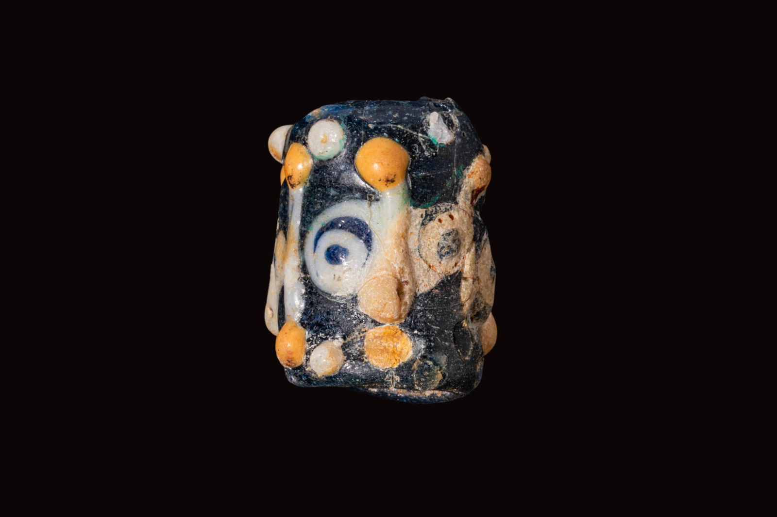 RARE PHOENICIAN GLASS FACE BEAD: Ca. 500 - 300 BC. A blue glass bead of cylindrical form with three faces spaced around the circumference, each with a prominent nose and eyes with blue pupils, the edges set with rows of alternating w
