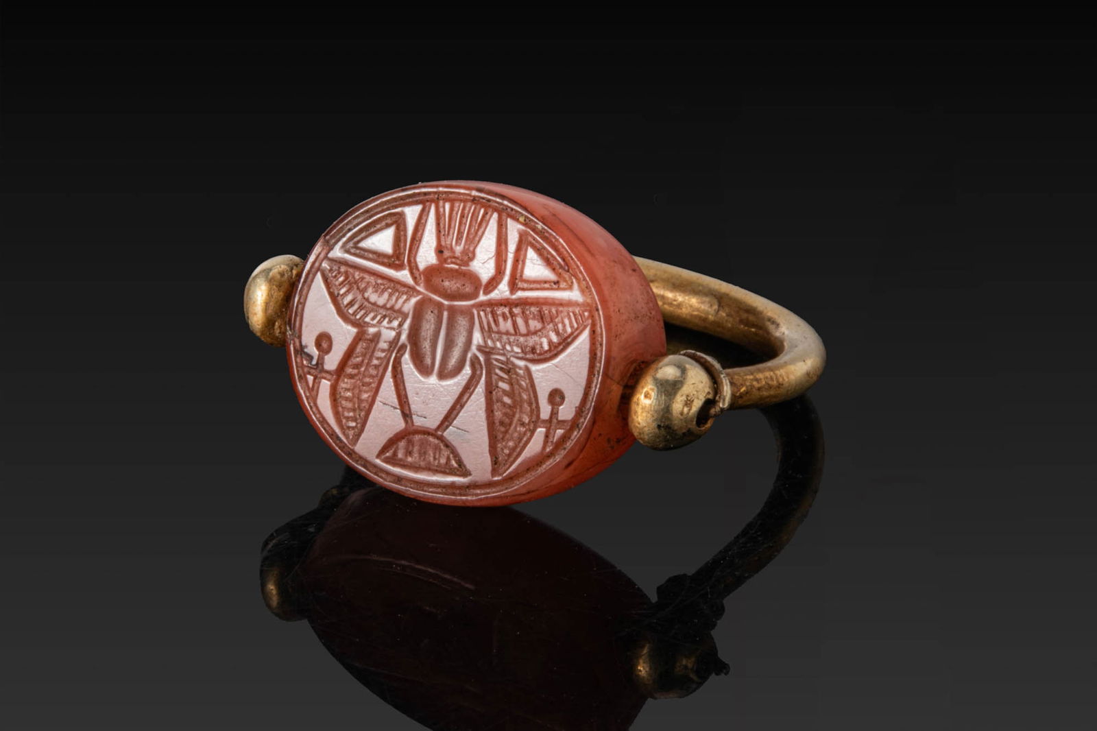 PHOENICIAN HIGH KARAT GOLD SWIVEL RING WITH CARNELIAN SCARAB WITH WINGED SCARAB: Ca. 600 - 400 BC. A gold swivel ring with a rounded hoop, fitted at the shoulders with wire-wound pivots and globular terminals supporting a carnelian scarab engraved with a winged scarab with outspre