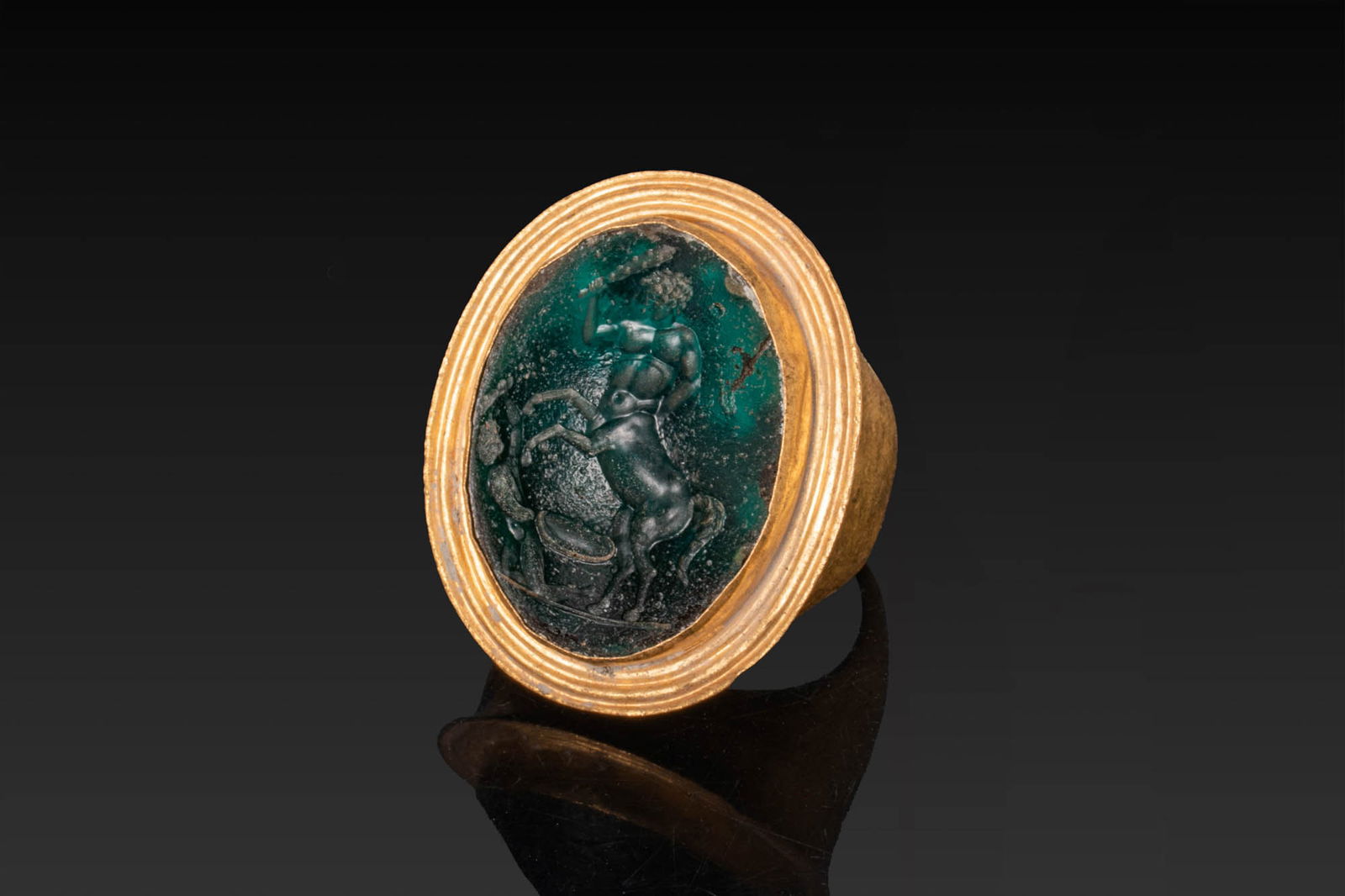 RARE ROMAN GREEN GLASS INTAGLIO OF CENTAUROMACHY IN HIGH KARAT GOLD RING: Ca. 100 BC - AD 100. A green glass intaglio engraved with a centauromachy, depicting a centaur rearing above a fallen figure with a shield, set in a possibly later gold ring with a broad oval bezel fo
