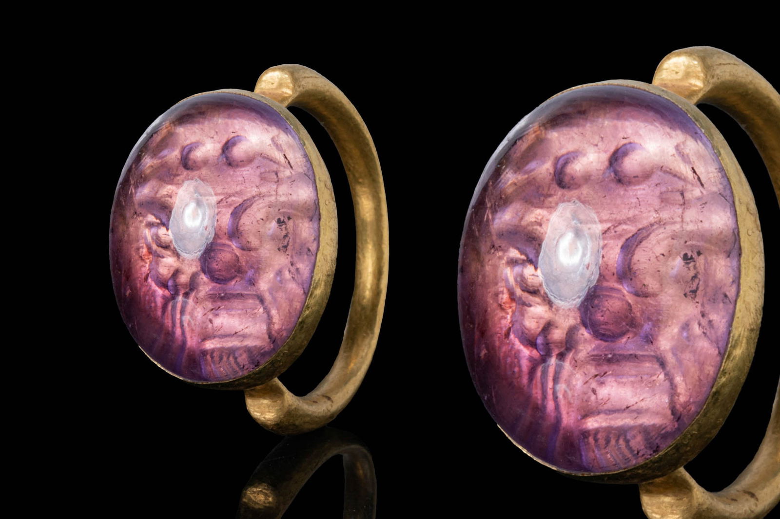 ROMAN AMETHYST HOLOGRAPHIC INTAGLIO OF A THEATRICAL MASK IN HIGH KARAT GOLD RING: Ca. AD 1 - 50. A convex amethyst holographic intaglio engraved with a theatrical mask shown frontally, with prominent eyes, arched brows and a short beard. Mounted in a gold ring with a slender hoop o
