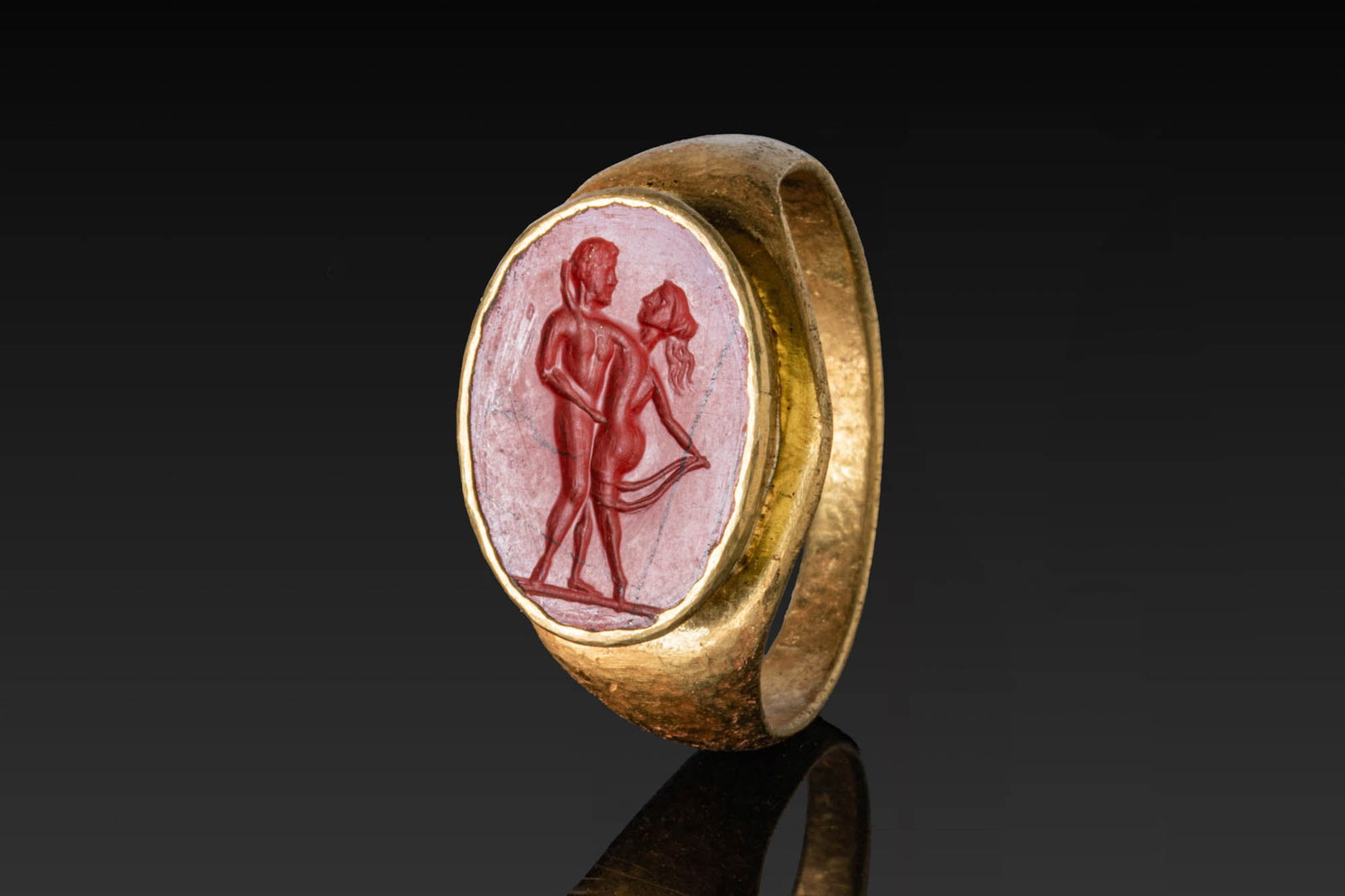 ROMAN RED EROTIC JASPER INTAGLIO OF A NUDE COUPLE IN HIGH KARAT GOLD RING: Ca. AD 300. A red jasper intaglio engraved with a nude couple standing face to face on a groundline. The ingalio is set in a possibly later high karat gold ring with a broad, flat-section hoop widenin