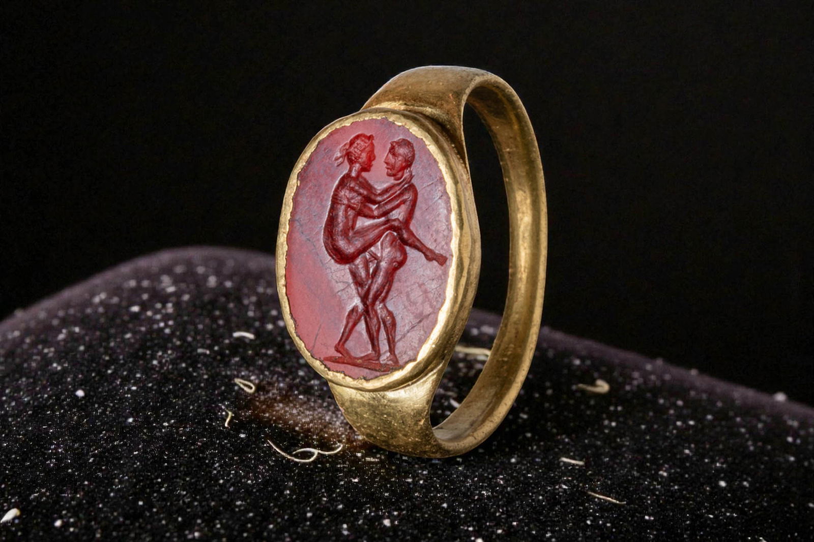 ROMAN CARNELIAN INTAGLIO OF AN EROTIC SCENE IN HIGH KARAT GOLD RING: Ca. 100 BC - AD 50. An oval carnelian intaglio engraved with an erotic scene, with a nude couple standing in profile, the male figure on a short groundline and the female with one leg raised against h