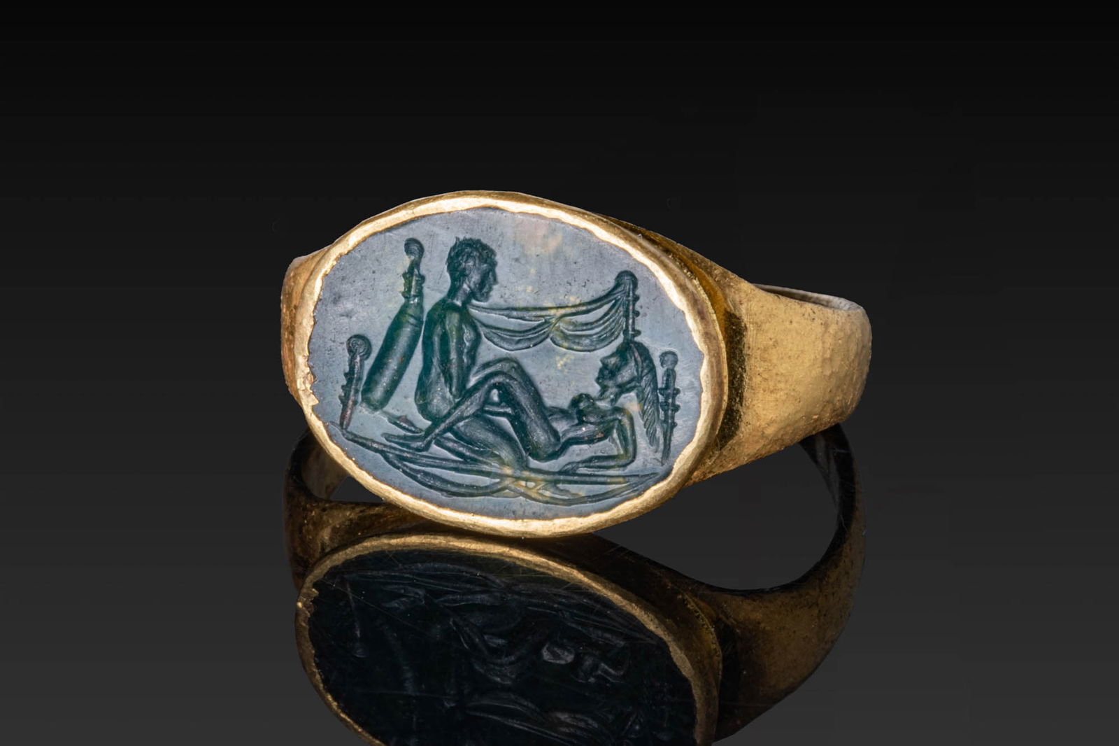 ROMAN GREEN JASPER INTAGLIO OF EROTIC SCENE IN HIGH KARAT GOLD RING: Ca. AD 1 - 300. A green jasper intaglio engraved with a nude couple on a couch beneath a swagged curtain, flanked by turned bedposts, set in a possibly later gold ring. The intaglio is secured in a ta