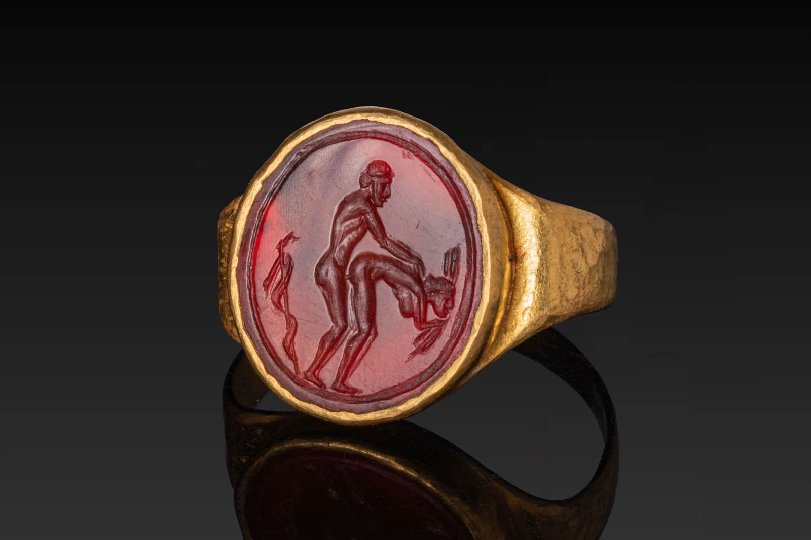 ROMAN (or later) CARNELIAN INTAGLIO OF AN EROTIC SCENE IN HIGH KARAT GOLD RING: Ca. AD 1 - 300 (or later). A carnelian intaglio engraved with an erotic scene, showing a nude male figure standing with one arm extended towards a partner bent forward, with a staff behind. Set in a p