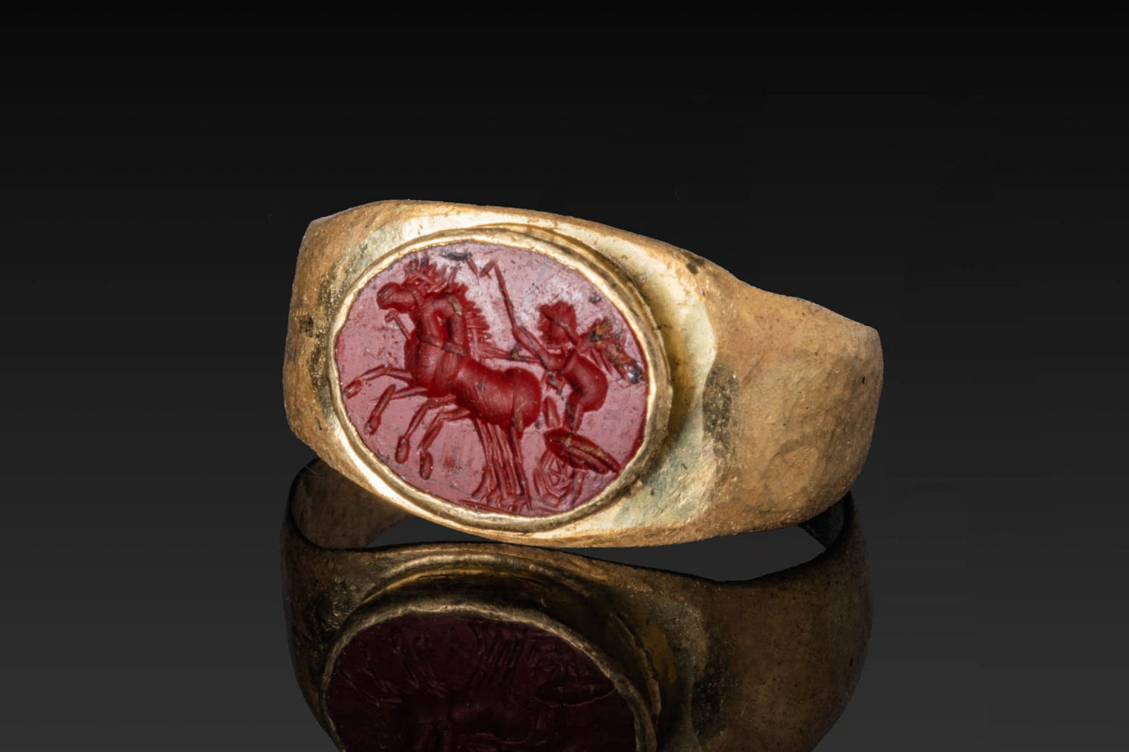 ROMAN RED JASPER INTAGLIO OF WINGED EROS DRIVING BIGA IN HIGH KARAT GOLD RING: Ca. AD 50 - 200. A red jasper intaglio engraved with winged Eros standing in a biga, holding the reins, with two horses in profile before him, set in a possibly later gold ring with a broad hoop of fl