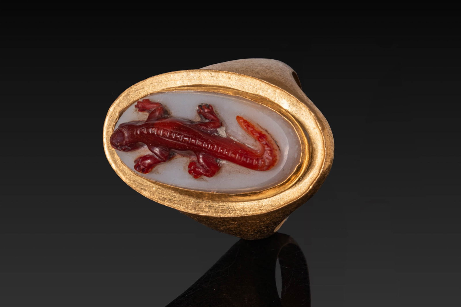 ROMAN AGATE CAMEO OF LIZARD IN HIGH KARAT HIGH KARAT GOLD RING: Ca. AD 1 - 300. An agate cameo carved in relief with a lizard, shown with splayed legs and a curling tail, modelled in the upper red layer against the pale ground, set in a possibly later high-karat g