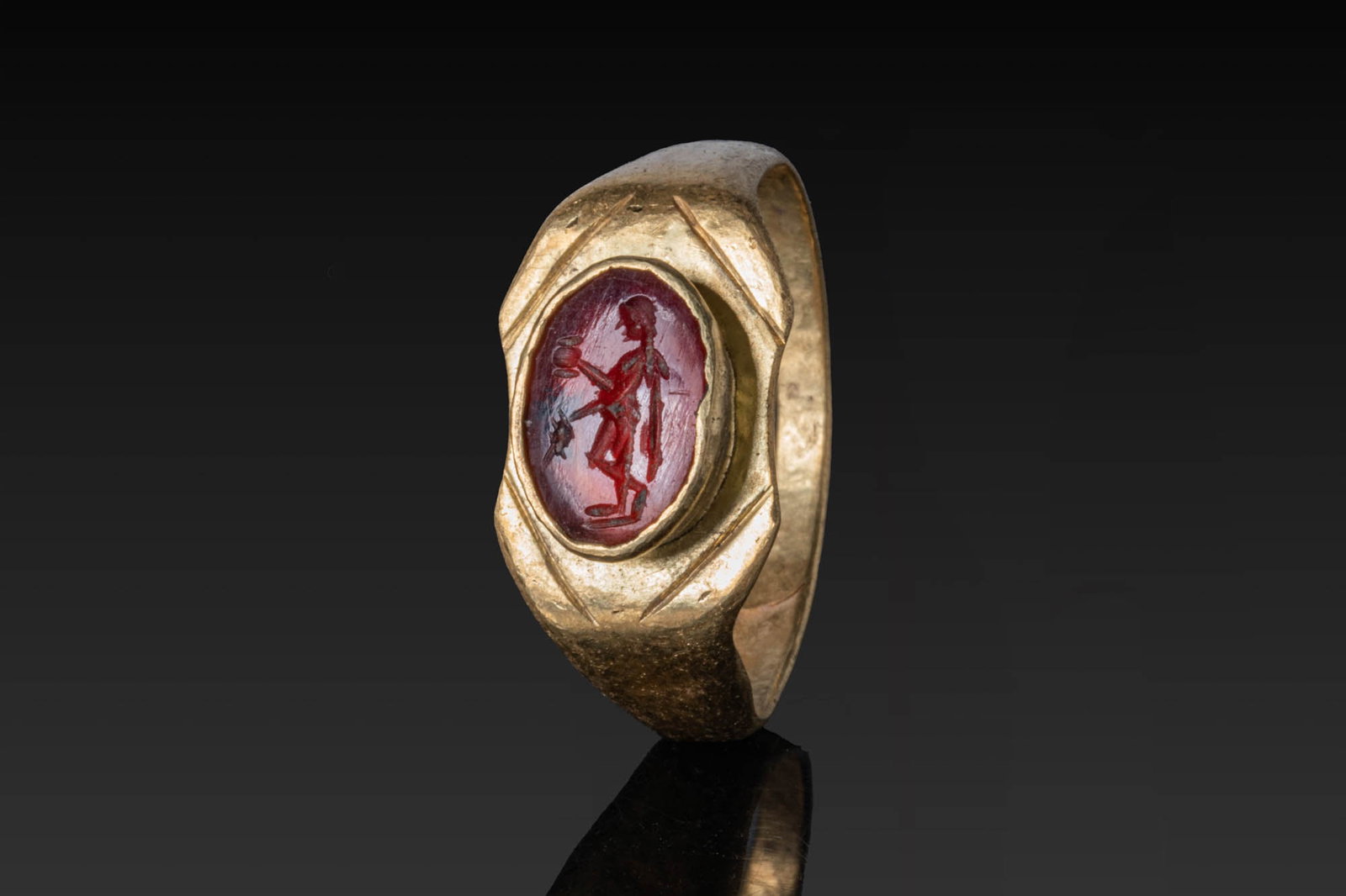 ROMAN RED GARNET INTAGLIO OF MERCURY IN HIGH KARAT GOLD RING: Ca. AD 1 - 300. A red garnet intaglio engraved with Mercury standing in profile, holding a staff, mounted in a possibly later gold ring with an octagonal bezel plate and raised oval collet, the facete