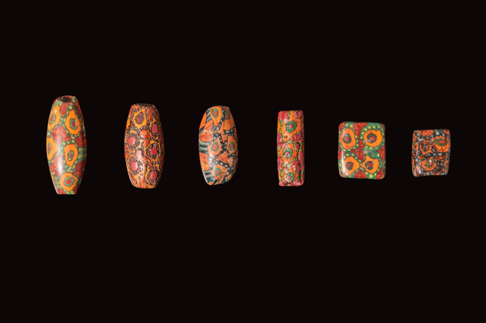 COLLECTION OF ROMAN MILLEFIORI GLASS BEADS: Ca. 100 BC - AD 100. A collection of six millefiori glass beads, each with polychrome mosaic decoration in red, orange, green and black; including barrel-shaped, cylindrical, and flattened rectangular