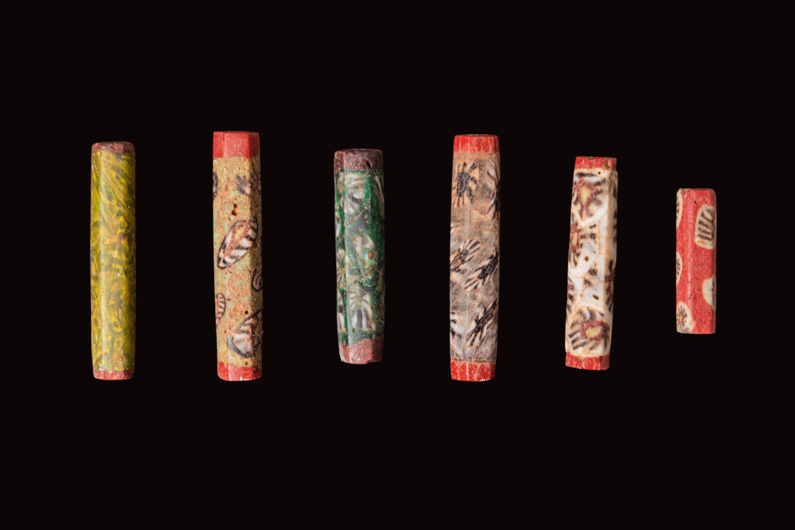 COLLECTION OF ROMAN MOSAIC GLASS CYLINDRICAL BEADS: Ca. 100 BC - AD 100. A collection of six faceted beads, each of long cylindrical form, in polychrome mosaic glass in red, yellow, green and cream, with marbled patterning. For similar see: MET Museum