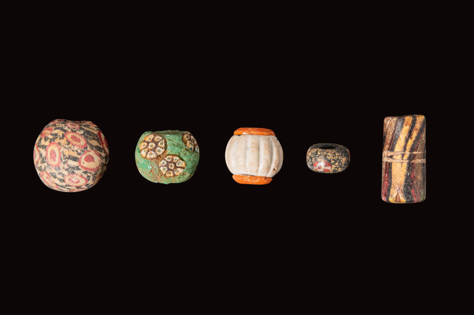 COLLECTION OF GRAECO-PHOENICIAN GLASS BEADS: Ca. 300 BC - AD 200. A collection of five glass beads, including, from left to right: a large globular bead with red circles on a white and black ground; a green barrel-shaped bead with seven-petalled