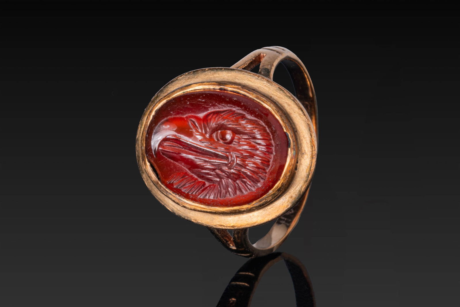 ROMAN CARNELIAN INTAGLIO OF AN EAGLE'S HEAD IN A VICTORIAN HIGH KARAT GOLD RING: Ca. AD 1 - 100. A carnelian intaglio engraved with an eagle?s head facing left, with a sharply hooked beak and a prominent, round eye. The oval stone is mounted in a Victorian gold ring with a broad b
