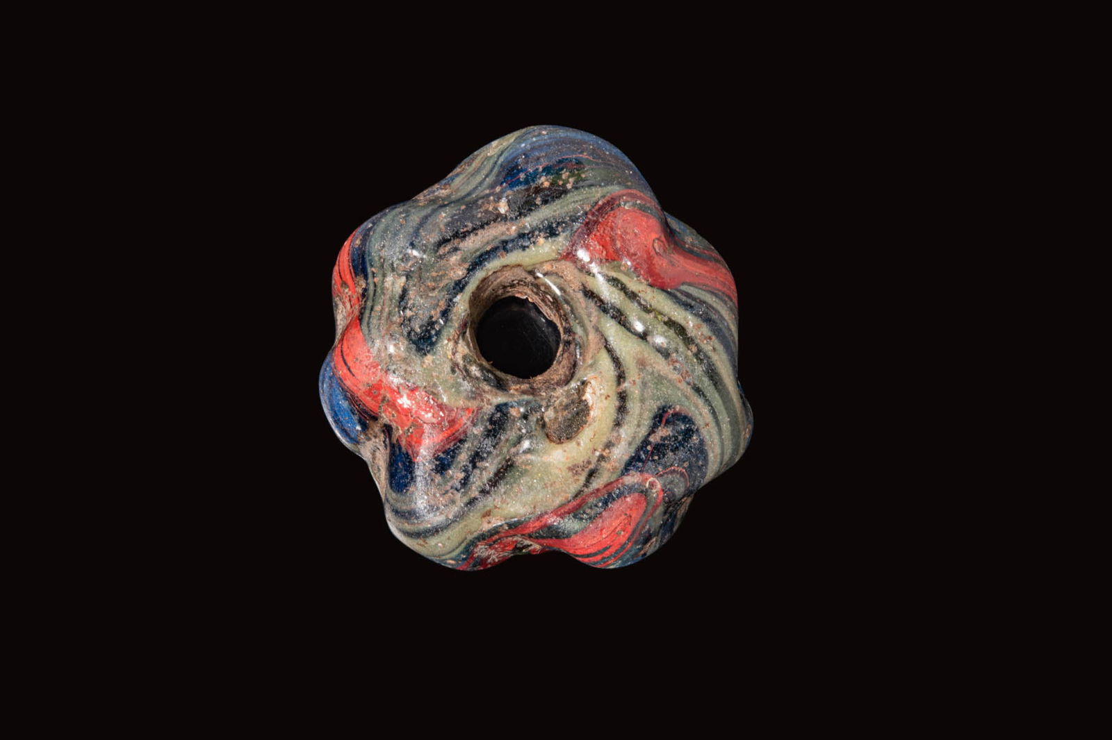 LARGE LATE ROMAN RIBBED GLASS BEAD: Ca. AD 300 - 600. A large ribbed glass bead of flattened annular form, decorated with marbled trails in cobalt blue, red, pale green and black. Size: 42mm x 15mm; Weight: 46g Provenance: Prince collec