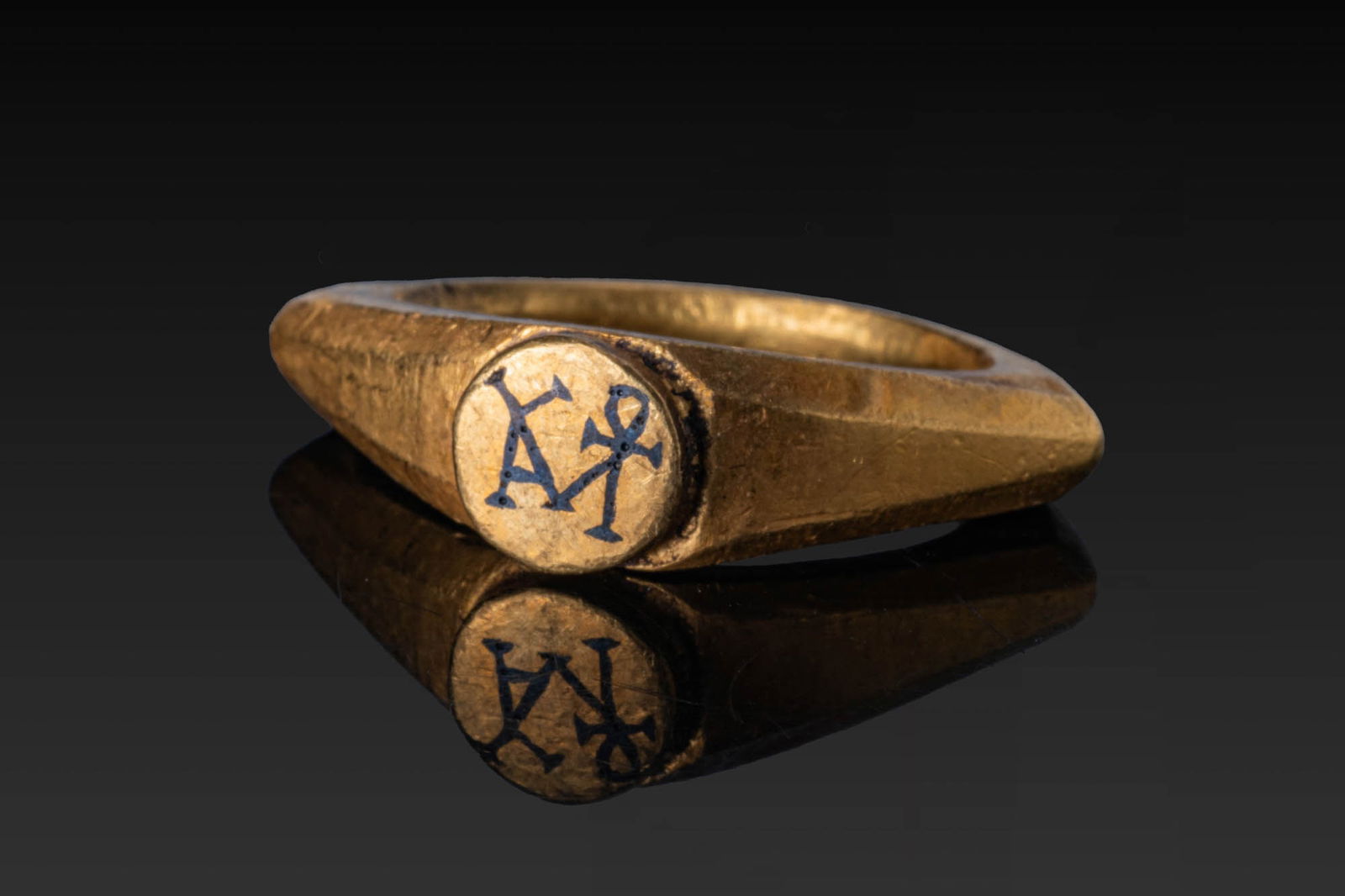 BYZANTINE HIGH KARAT GOLD RING WITH NIELLO MONOGRAM: Ca. AD 400 - 600. A gold ring with a faceted hoop, widening at the shoulders to support a circular bezel plate inlaid in niello with a monogram. For similar see: The British Museum, Museum number OA.7