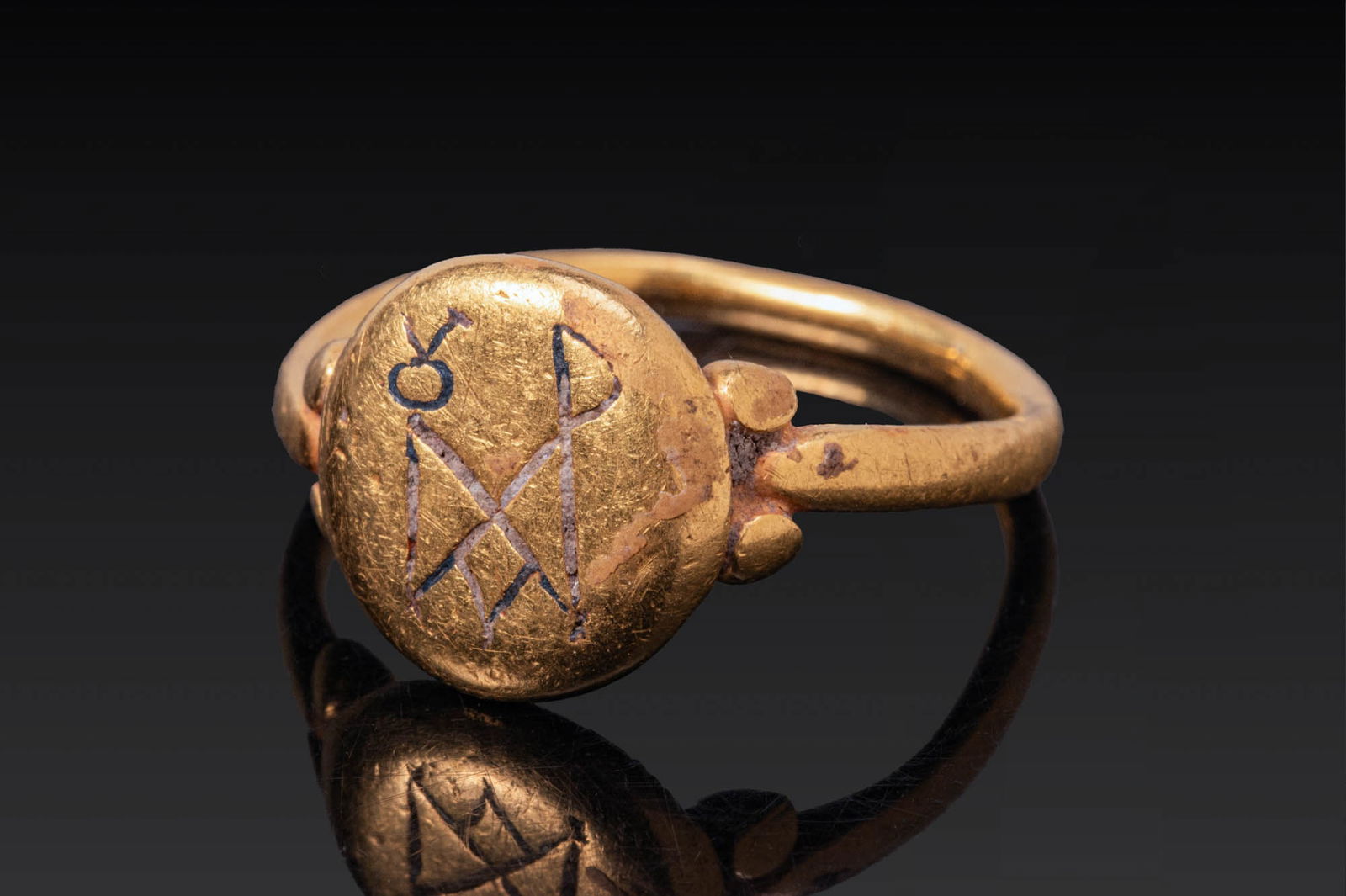 BYZANTINE HIGH KARAT GOLD AND NIELLO RING WITH MONOGRAM: Ca. AD 600. A gold ring with a plain, slender hoop and an attached circular bezel plate, the shoulders with pairs of applied pellets. The bezel is inlaid in niello with a complex interlaced monogram.