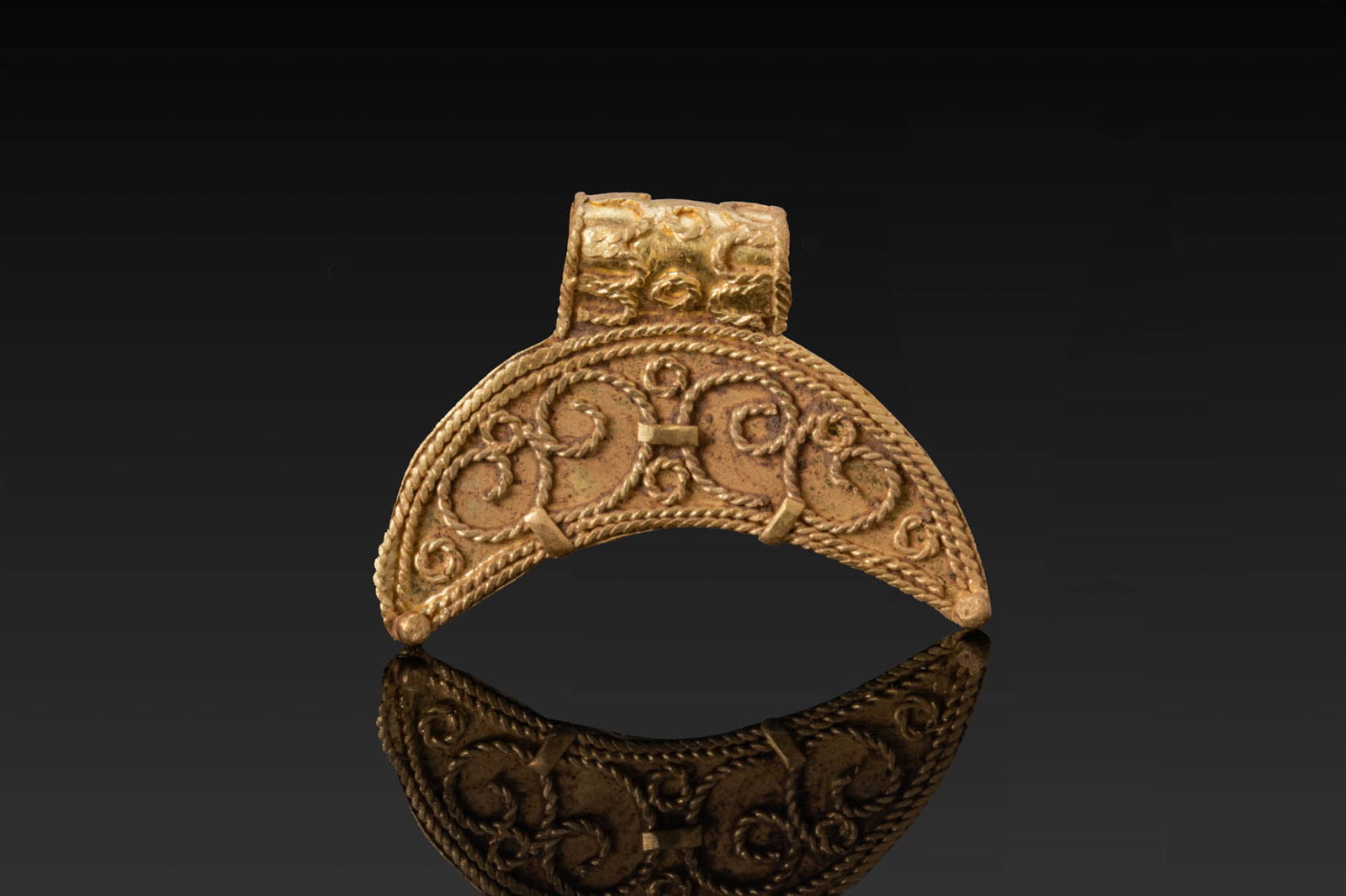 VIKING PERIOD HIGH KARAT GOLD ROPEWORK LUNAR PENDANT: Ca. AD 1000 - 1200. A gold crescent pendant with twisted wire borders. The obverse is decorated with paired scrolls and small circles. A suspension loop is soldered at the apex. The reverse is plain.
