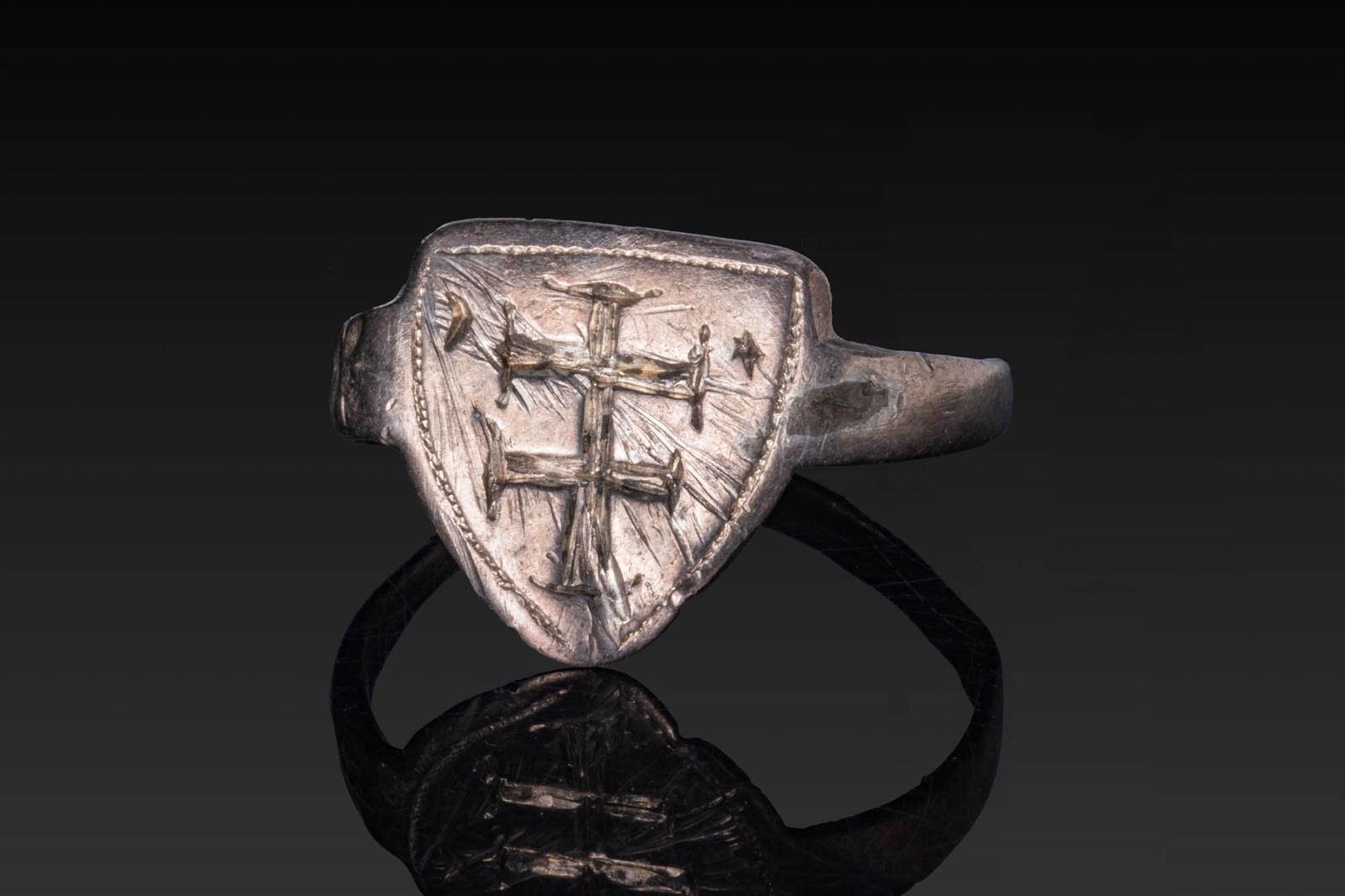 CRUSADER PERIOD SILVER RING WITH CROSS: Ca. AD 1100 - 1300. A silver ring with a slender hoop expanding to a shield-shaped bezel plate engraved with a double-barred cross with flared terminals, set within a beaded border, flanked by a cresc