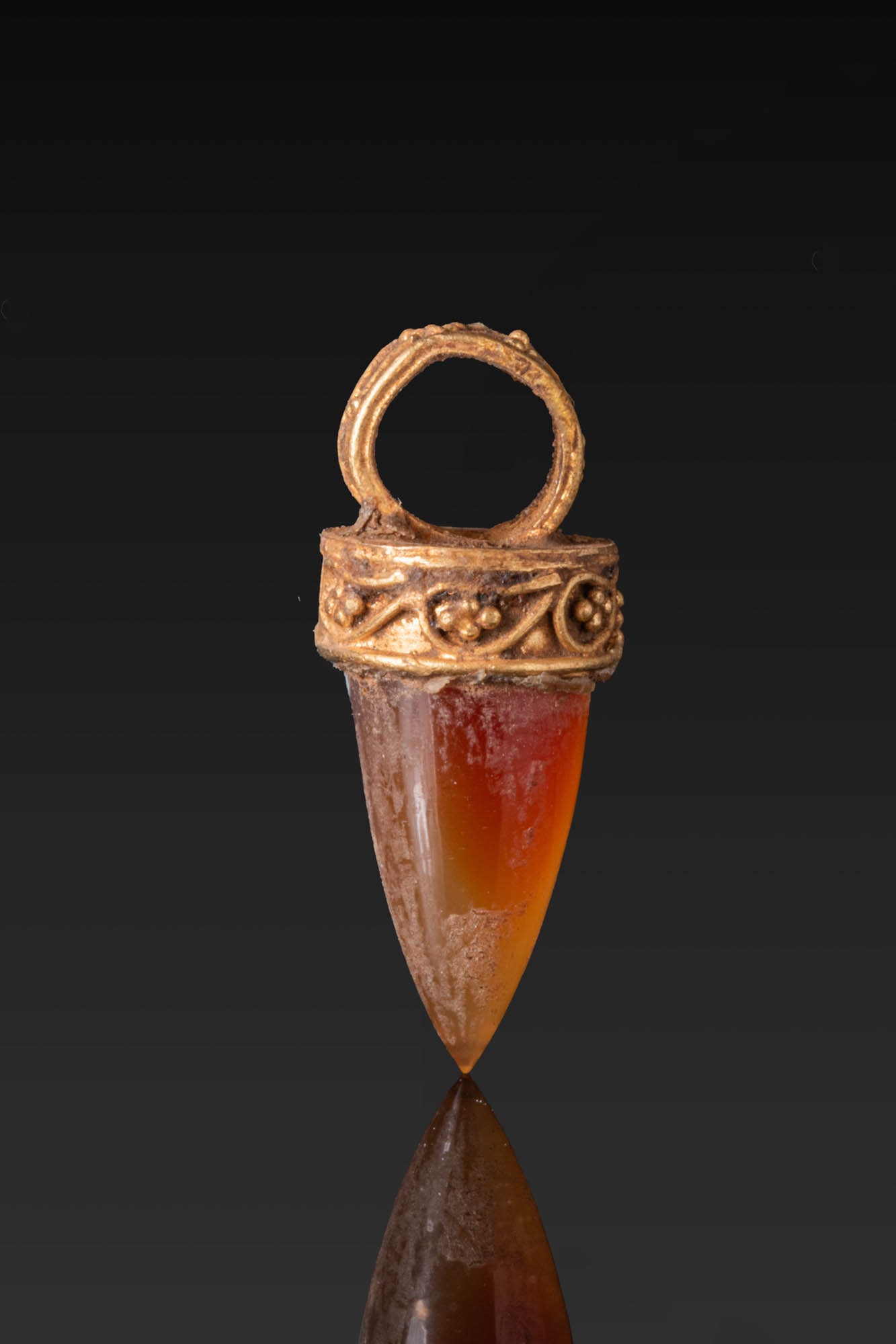 HELLENISTIC HIGH KARAT GOLD PENDANT WITH CARNELIAN: Ca. 300 - 100 BC. A gold pendant set with a tapered carnelian drop in a circular mount with a plain upper plate, the collar and suspension loop decorated with granulation and drop-shaped motifs in fin