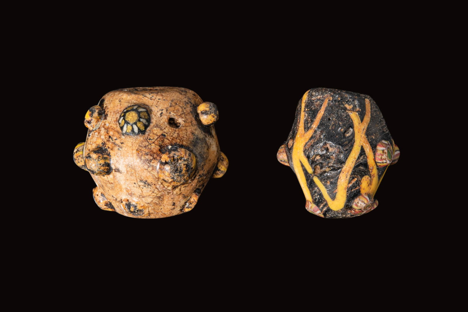 PAIR OF PHOENICIAN GLASS BEADS: Ca. 500 - 300 BC. A pair of glass biconical beads, from left to right: a squat bead with three rows of applied bosses decorated as rosettes; and a bead in dark glass with applied yellow trails and sma