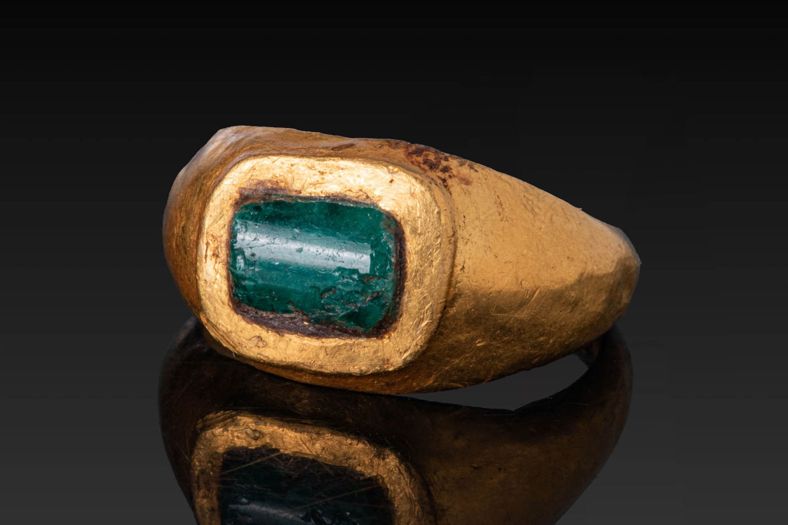 ROMAN HIGH KARAT GOLD RING WITH EMERALD BEZEL: Ca. AD 1 - 200. A gold ring with a broad tapering hoop of D-section and a rectangular bezel plate with rounded corners, set with an emerald. Size: D:14.88mm / US: 3 3/4 / UK: H; Weight: 3.9g Provenanc