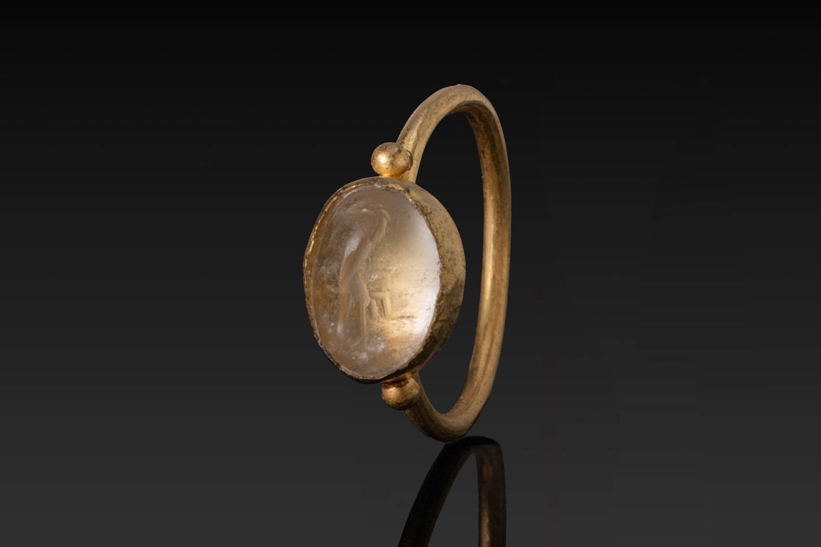 LATE ROMAN HIGH KARAT GOLD RING WITH CHALCEDONY INTAGLIO OF A STORK: Ca. AD 200 - 400. A gold ring with a slender hoop of rounded section and an oval bezel mount set with a chalcedony intaglio. Small globules are applied at the top and bottom of the setting. The intagl