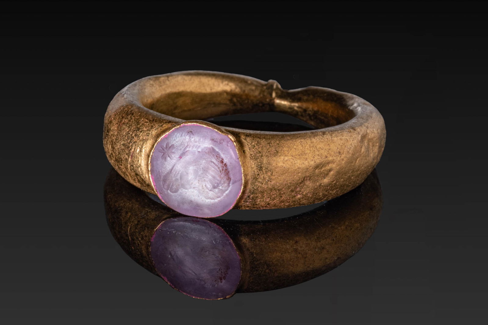 ROMAN AMETHYST INTAGLIO OF A RAM IN GOLD RING: Ca. AD 300 - 400. A circular amethyst intaglio engraved with the head of a ram in left profile, accompanied by an eight-pointed star above. Set in a possibly later gold ring with a round bezel setting