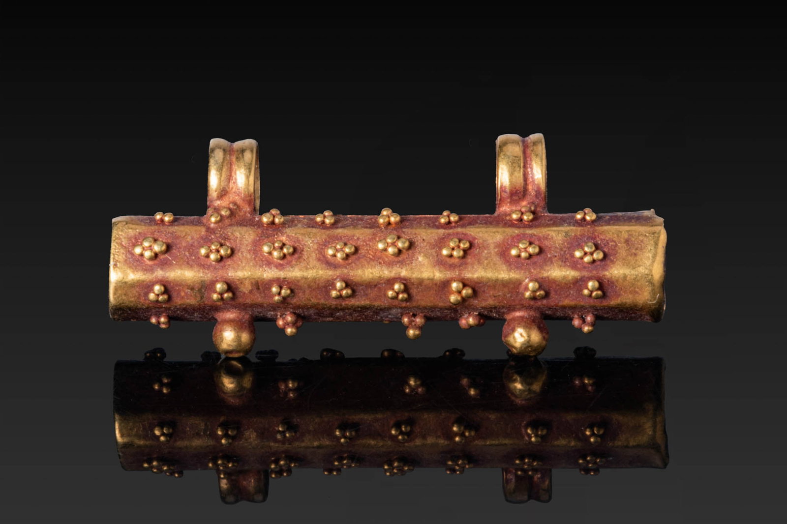 ROMAN HIGH KARAT GOLD SCROLL PENDANT WITH GRANULATION: Ca. AD 1 - 300. A gold tubular pendant with two suspension loops. The sides are decorated with clustered granules in rows, with two bosses on the underside. Size: 26mm x 10mm; Weight: 2.1g Provenance:
