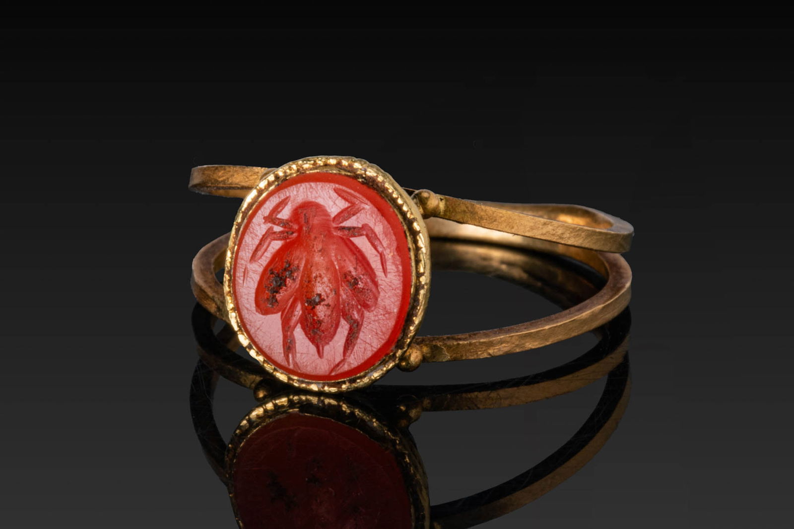 ROMAN CARNELIAN INTAGLIO OF A FLY IN HIGH KARAT GOLD RING WITH PELLETS: Ca. AD 1 - 300. A carnelian intaglio engraved with a fly shown from above. The stone is set within a narrow beaded frame and mounted in a gold ring with a paired hoop of slender bands with globular kn