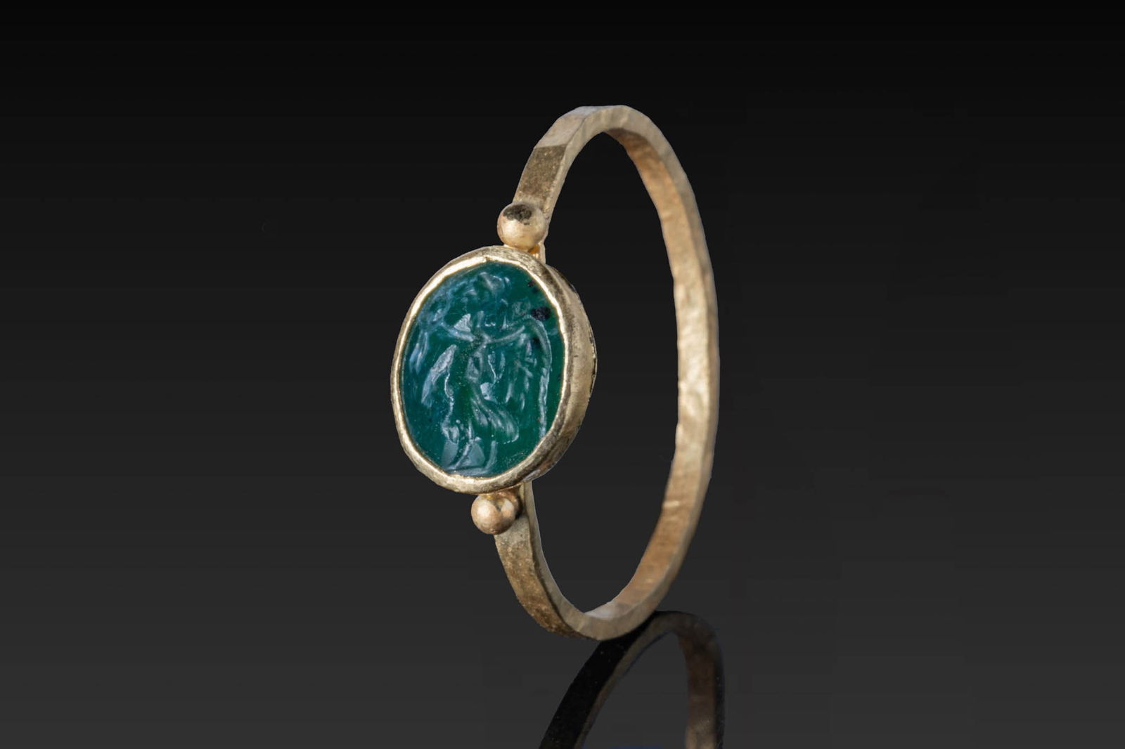 ROMAN EMERALD INTAGLIO OF WINGED VICTORY IN HIGH KARAT GOLD RING: Ca. AD 50 - 200. An oval emerald intaglio engraved with winged Victory, set in a possibly later gold ring. The bezel cell is flanked at the shoulders by small granules, on a slender hoop of faceted se
