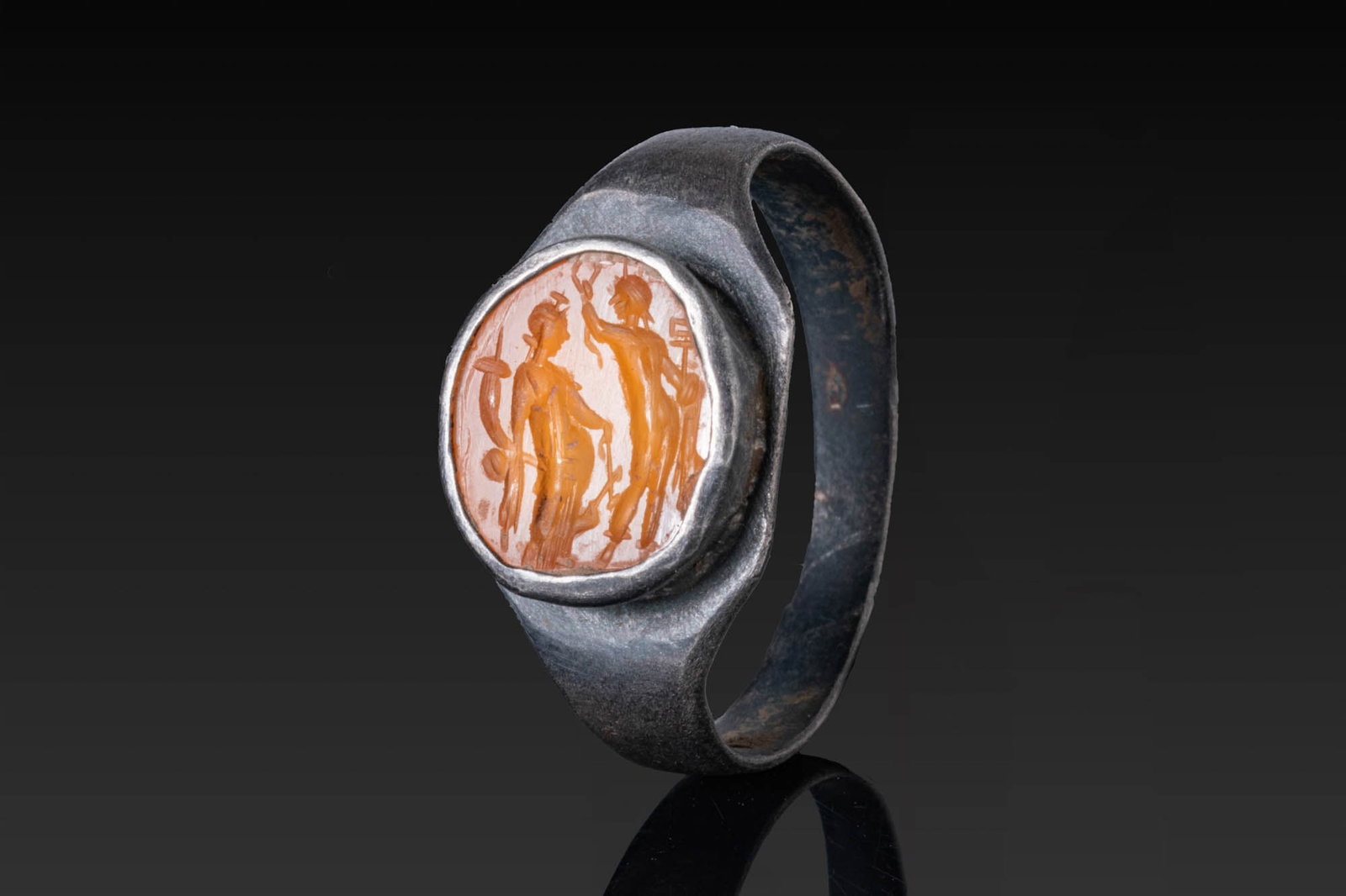 ROMAN CARNELIAN INTAGLIO OF ISIS FORTUNA AND ASCLEPIUS IN SILVER RING: Ca. AD 300. A carnelian intaglio engraved with Isis-Fortuna holding a cornucopia in the right hand and a rudder in the left, confronted by Asclepius with a staff and a serpent. Mounted in a possibly l